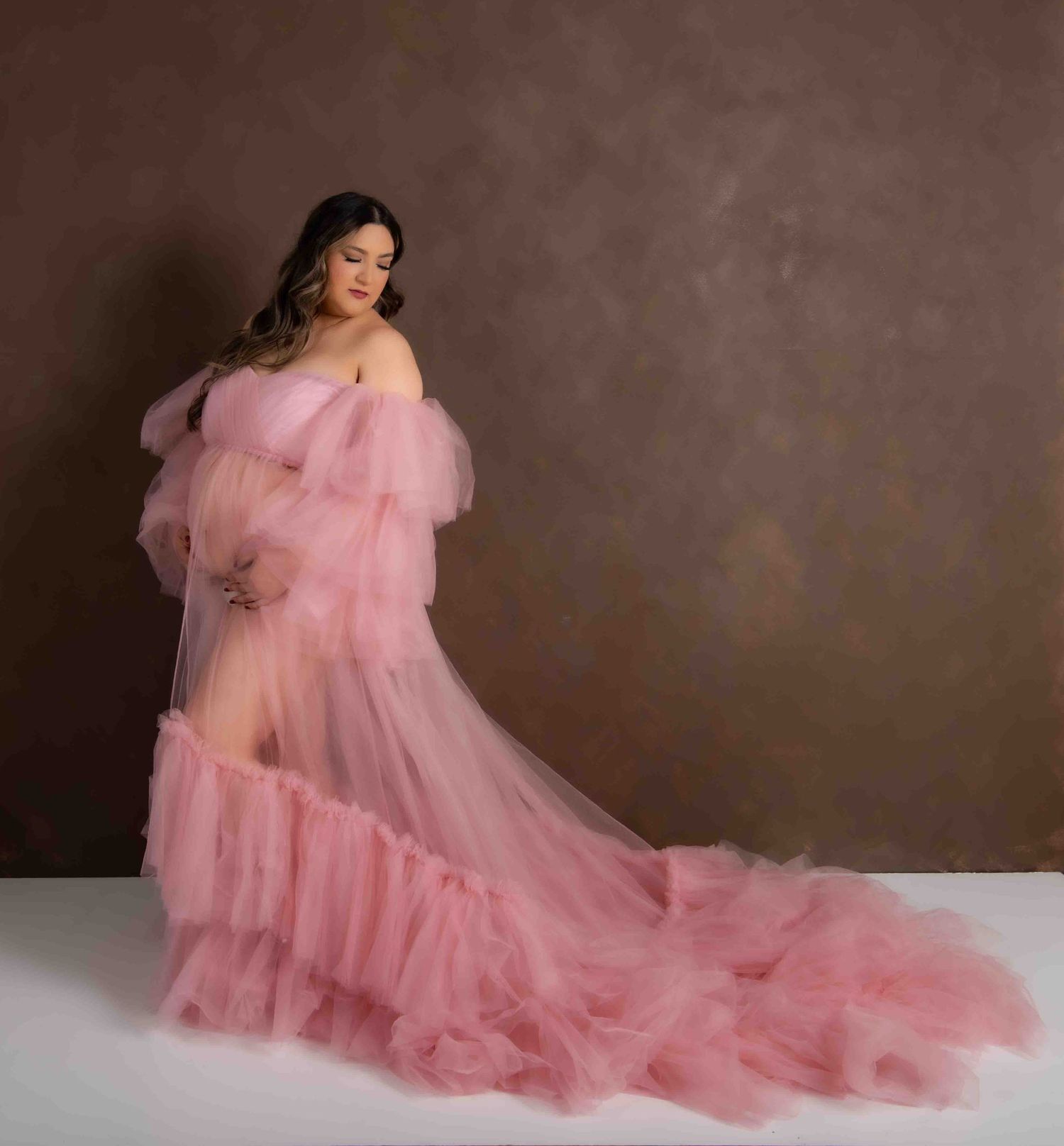 A pregnant woman poses elegantly in a flowing pink tulle maternity gown against a brown studio backdrop.