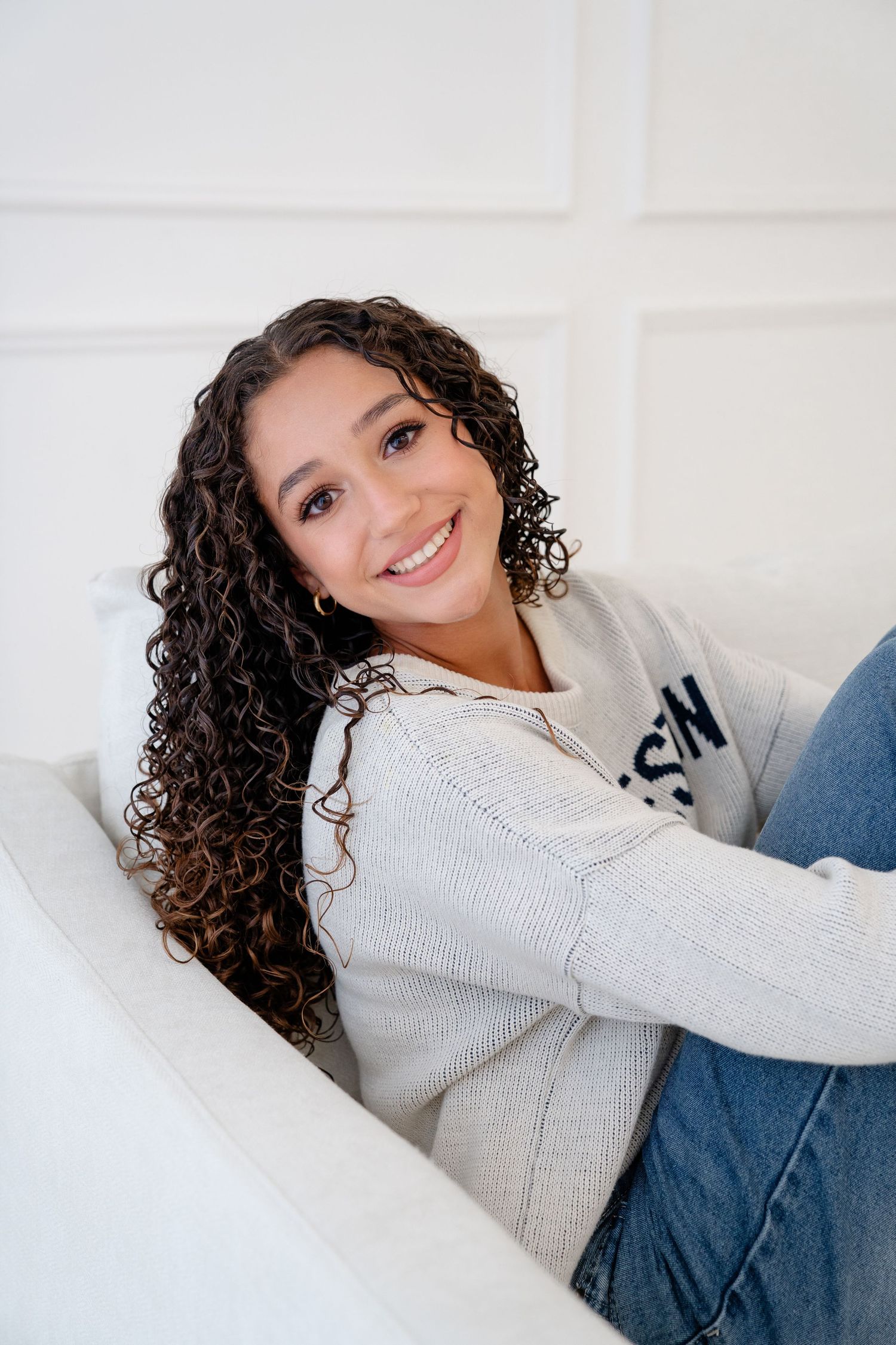 Young woman with curly hair smiling on white couch in gray hoodie and jeans.