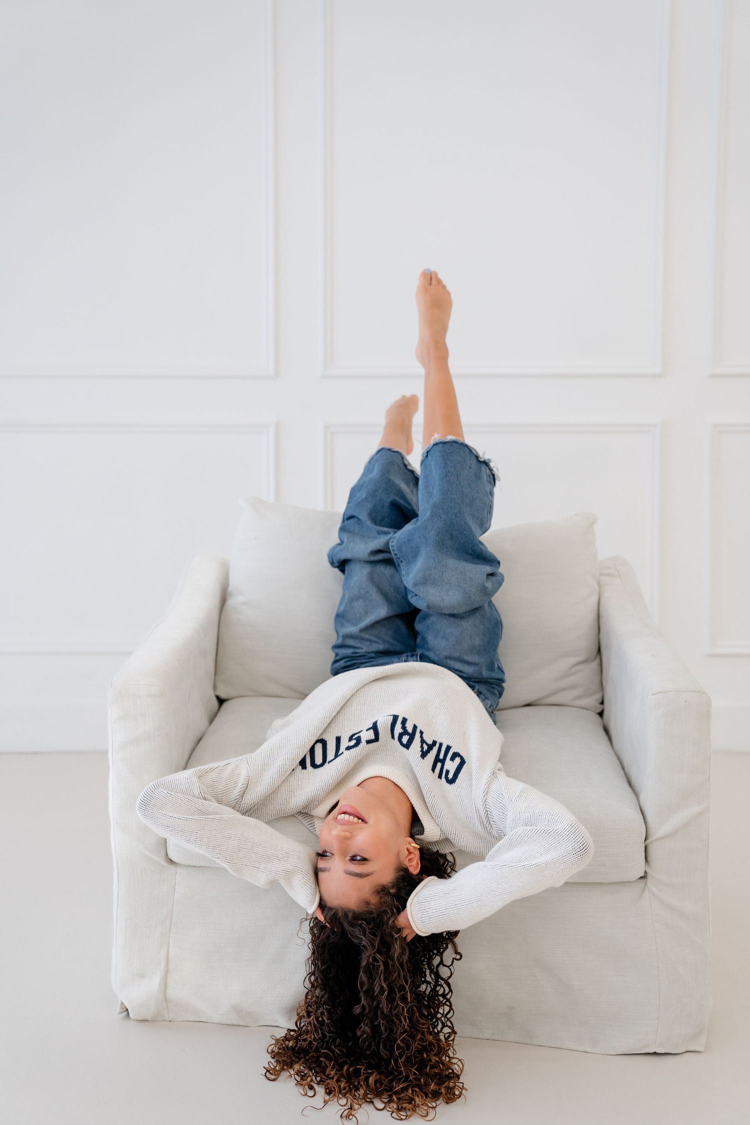 Playful child lying upside down on a white armchair, wearing jeans and a sweatshirt, laughing with legs in the air.
