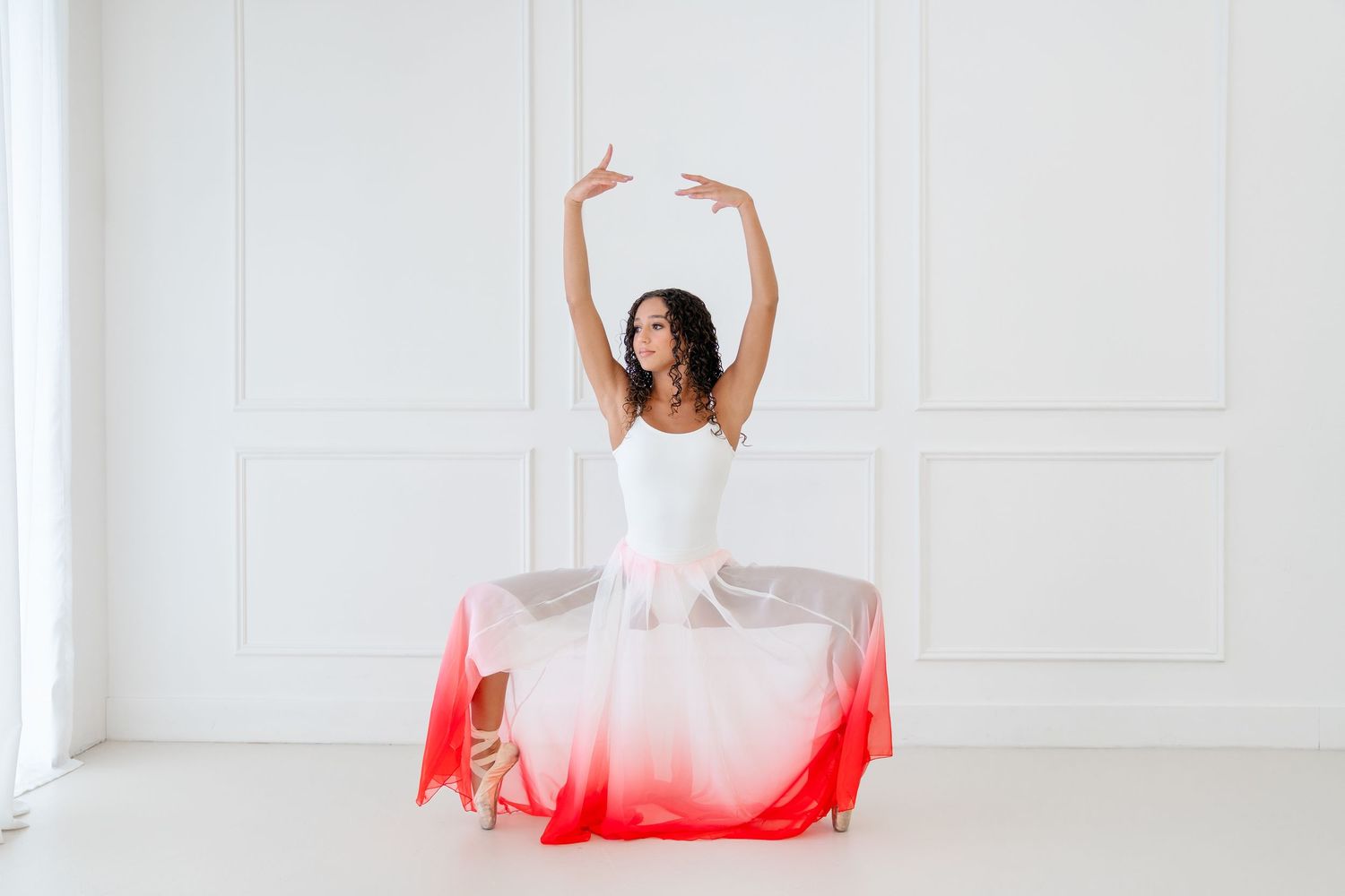Ballerina in white bodice and red ombre skirt performs splits on floor with arms raised in bright white studio.
