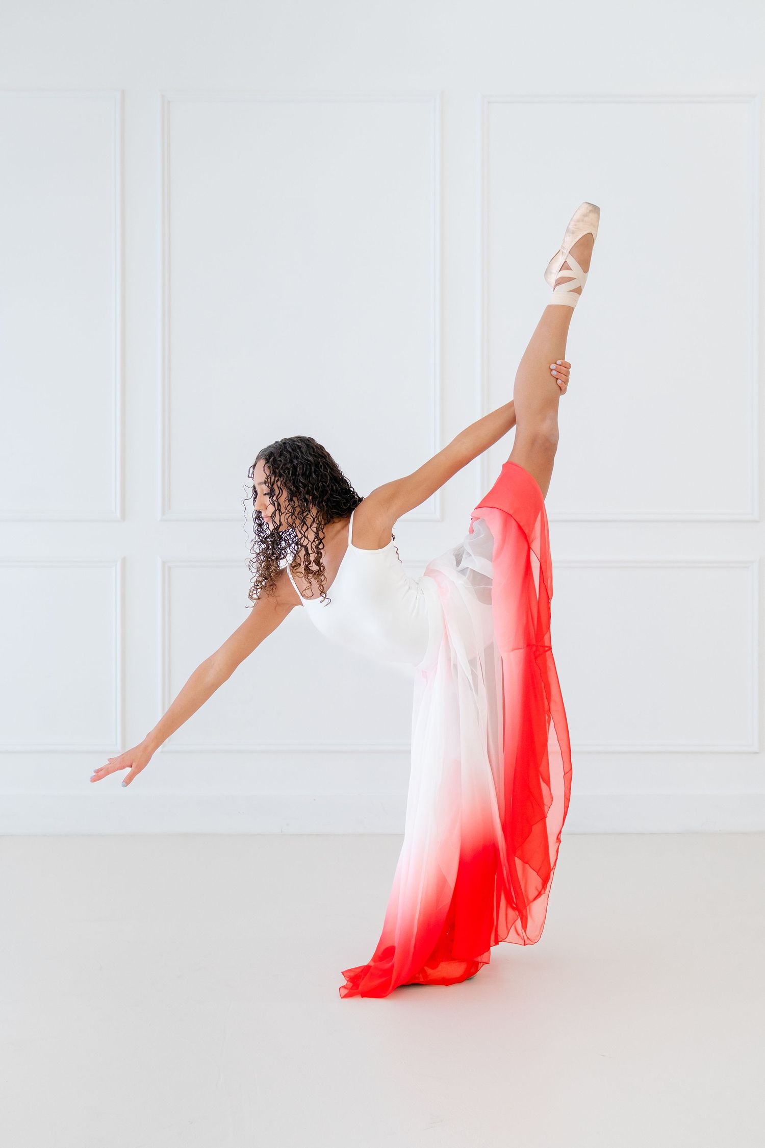 A ballerina in a white and red ombre dress performs an arabesque in pointe shoes against a white studio wall.