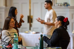 Two women conversing at a training workshop table with colleagues standing in background.