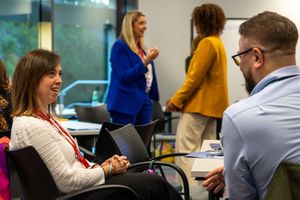 Professional woman laughs during business meeting while colleagues network in background at corporate event.