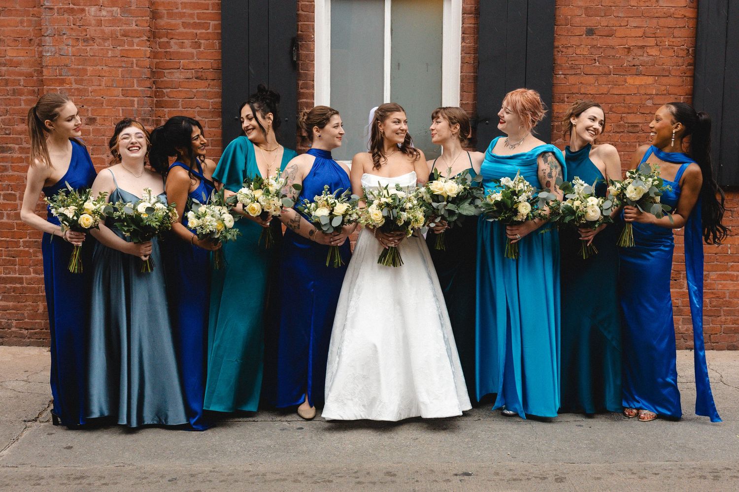 Bride in white gown stands with bridesmaids in blue and teal dresses holding bouquets outside brick building.