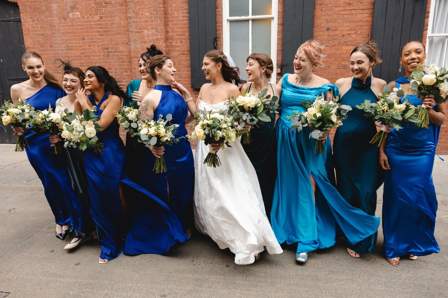 Bride in white gown walks joyfully with bridesmaids in blue and teal dresses holding white bouquets.