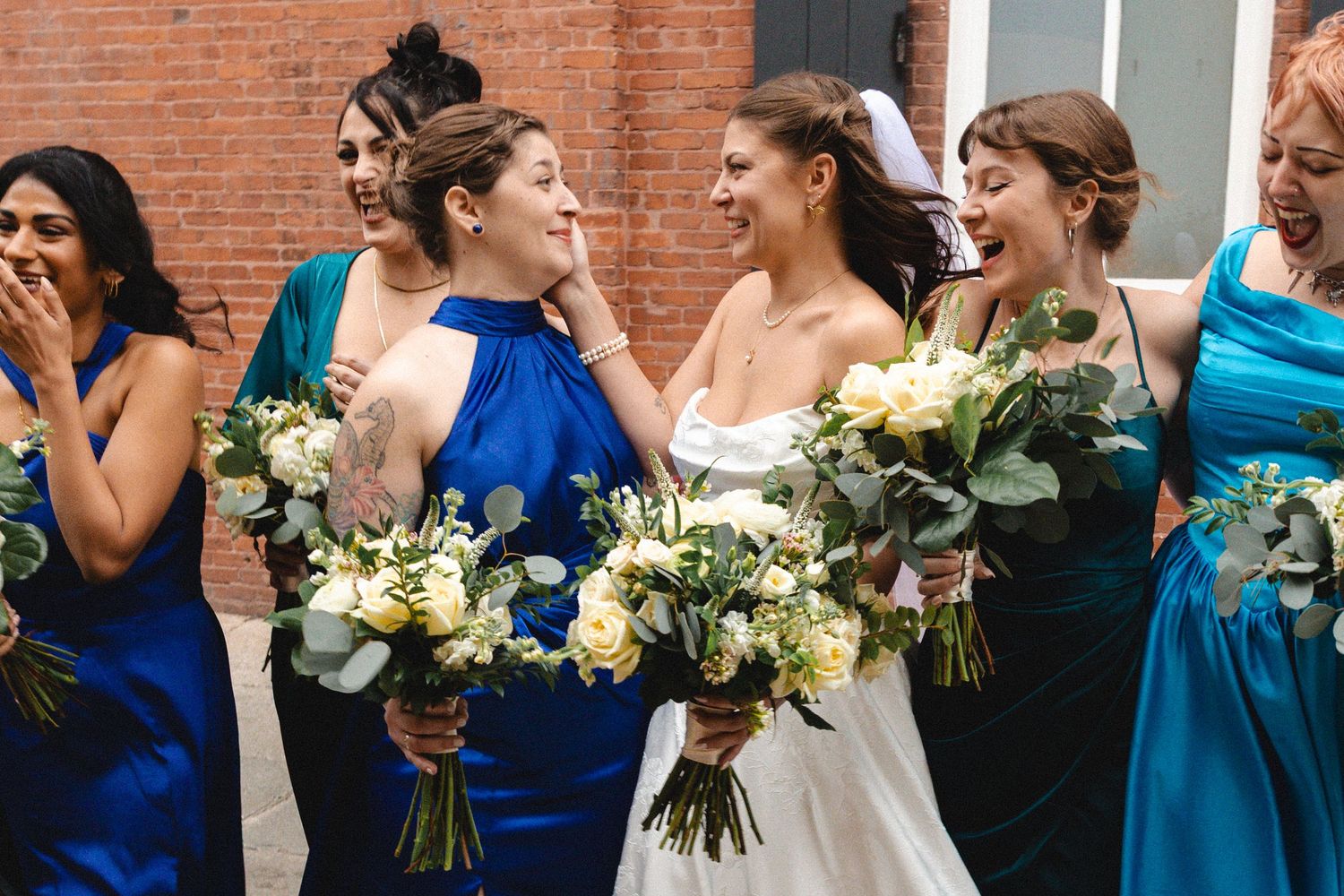 Bride and bridesmaids in blue dresses laughing together while holding white and green bouquets outdoors.