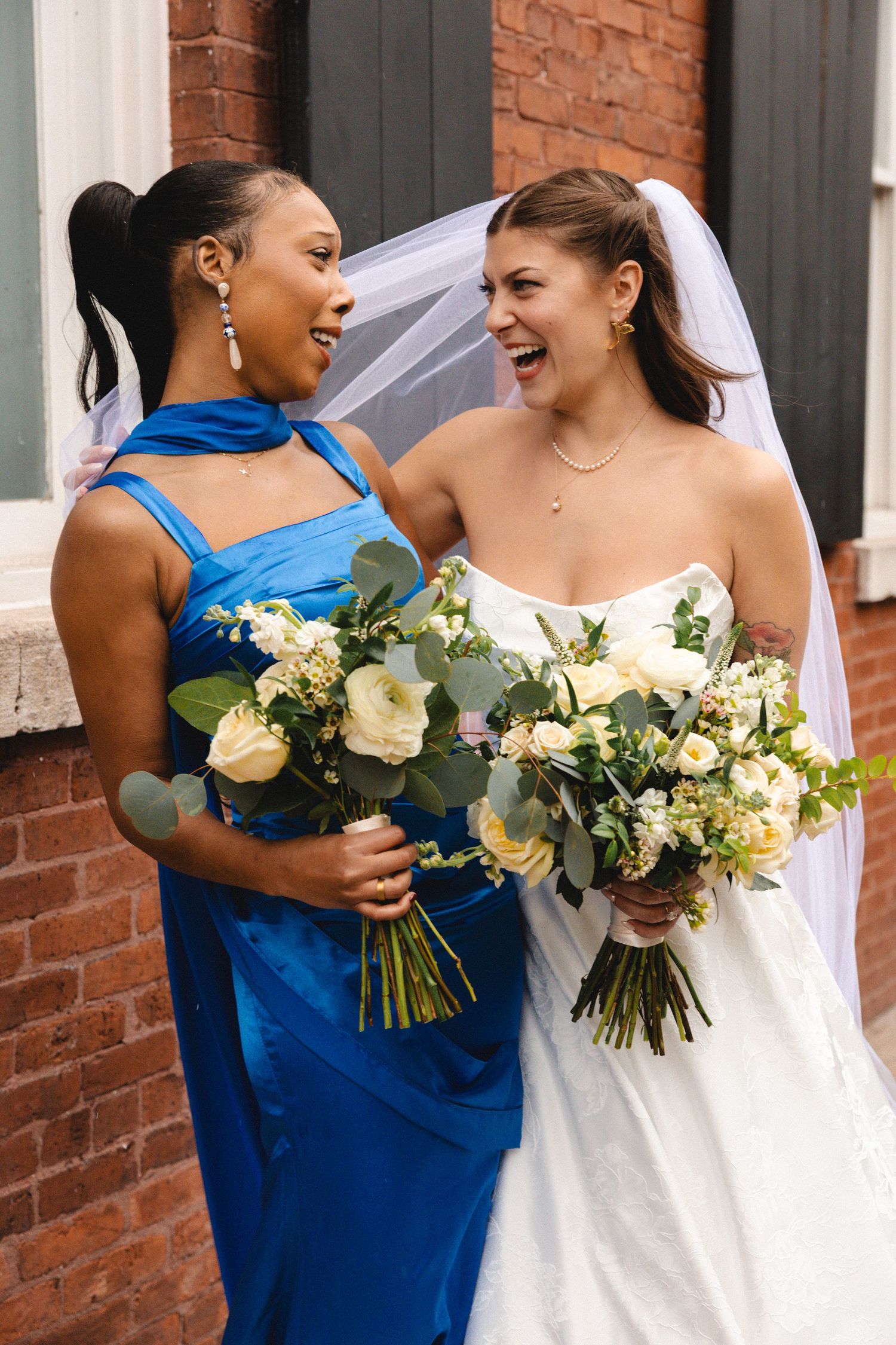 Bride in white gown and bridesmaid in blue dress laughing together while holding white floral bouquets outdoors.
