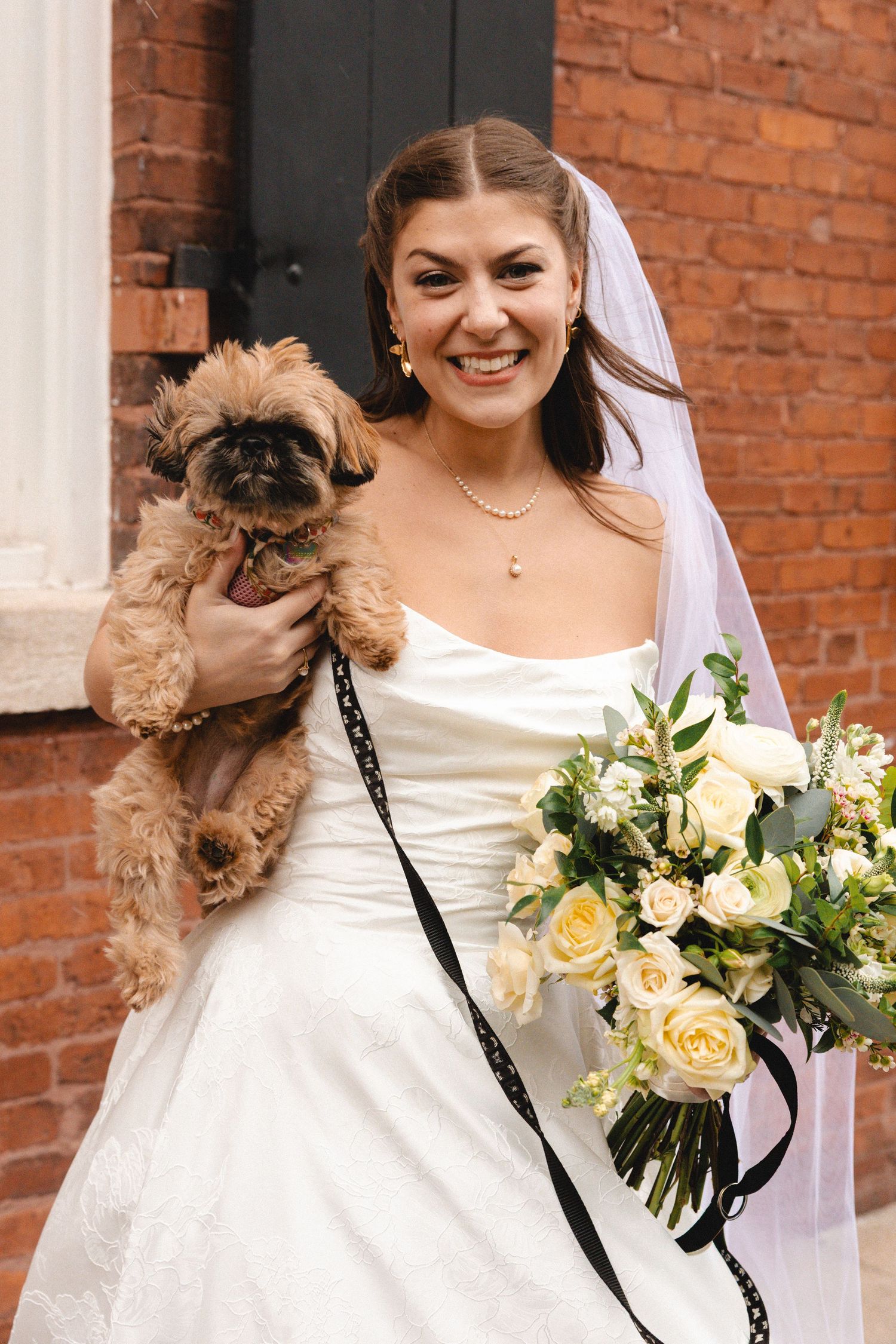 Smiling bride in white gown holding fluffy brown dog and white rose bouquet, standing by brick wall.