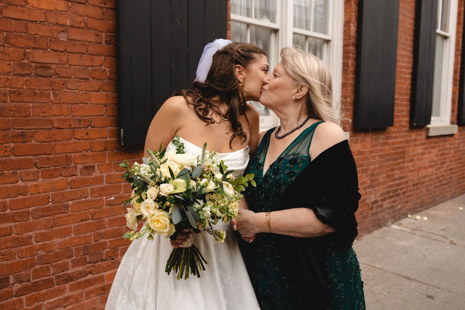 Bride in white gown kissing older woman in green dress, holding white floral bouquet, near brick wall.
