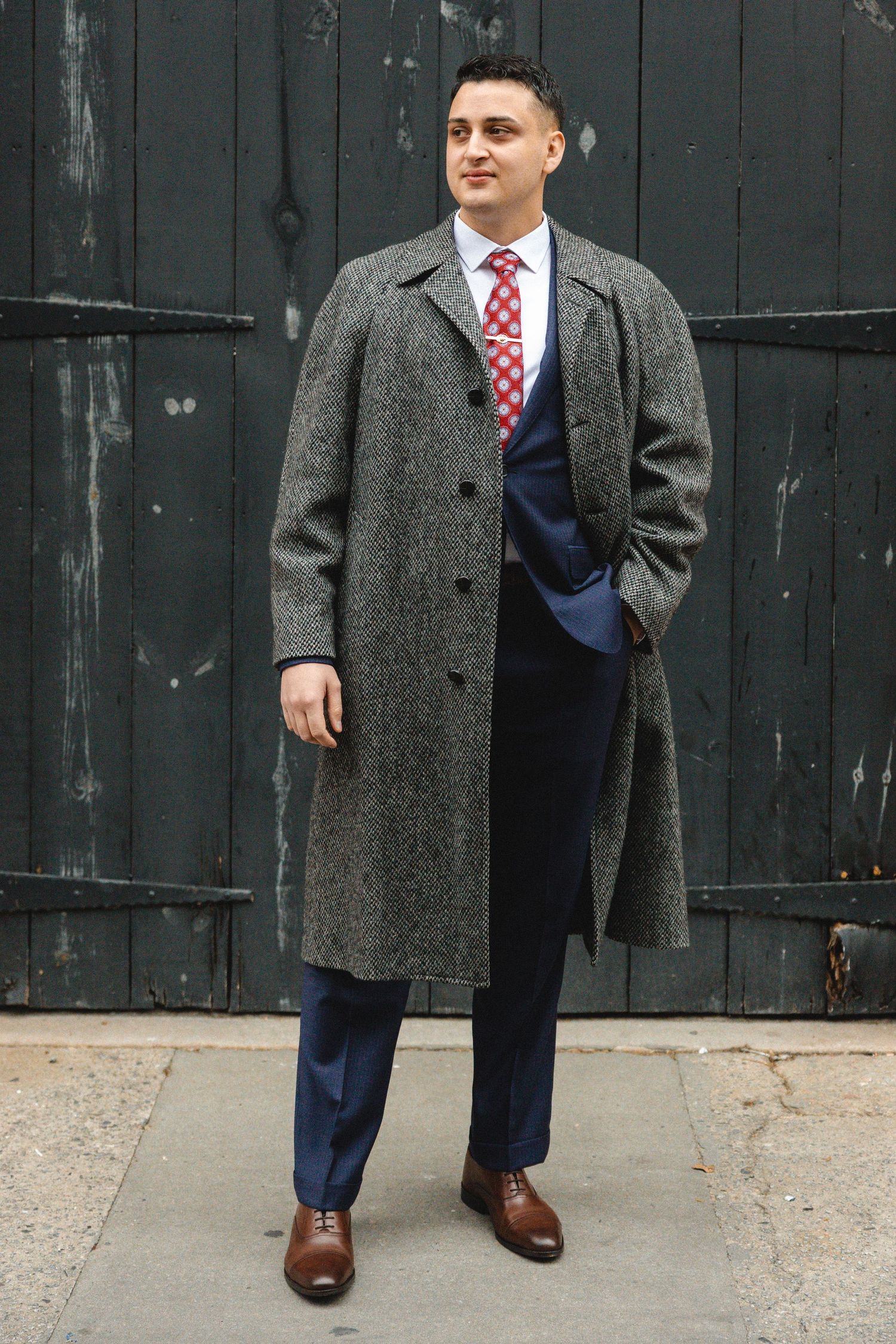 Man in gray tweed overcoat, navy suit, and red tie poses confidently on a sidewalk against a dark wooden backdrop.