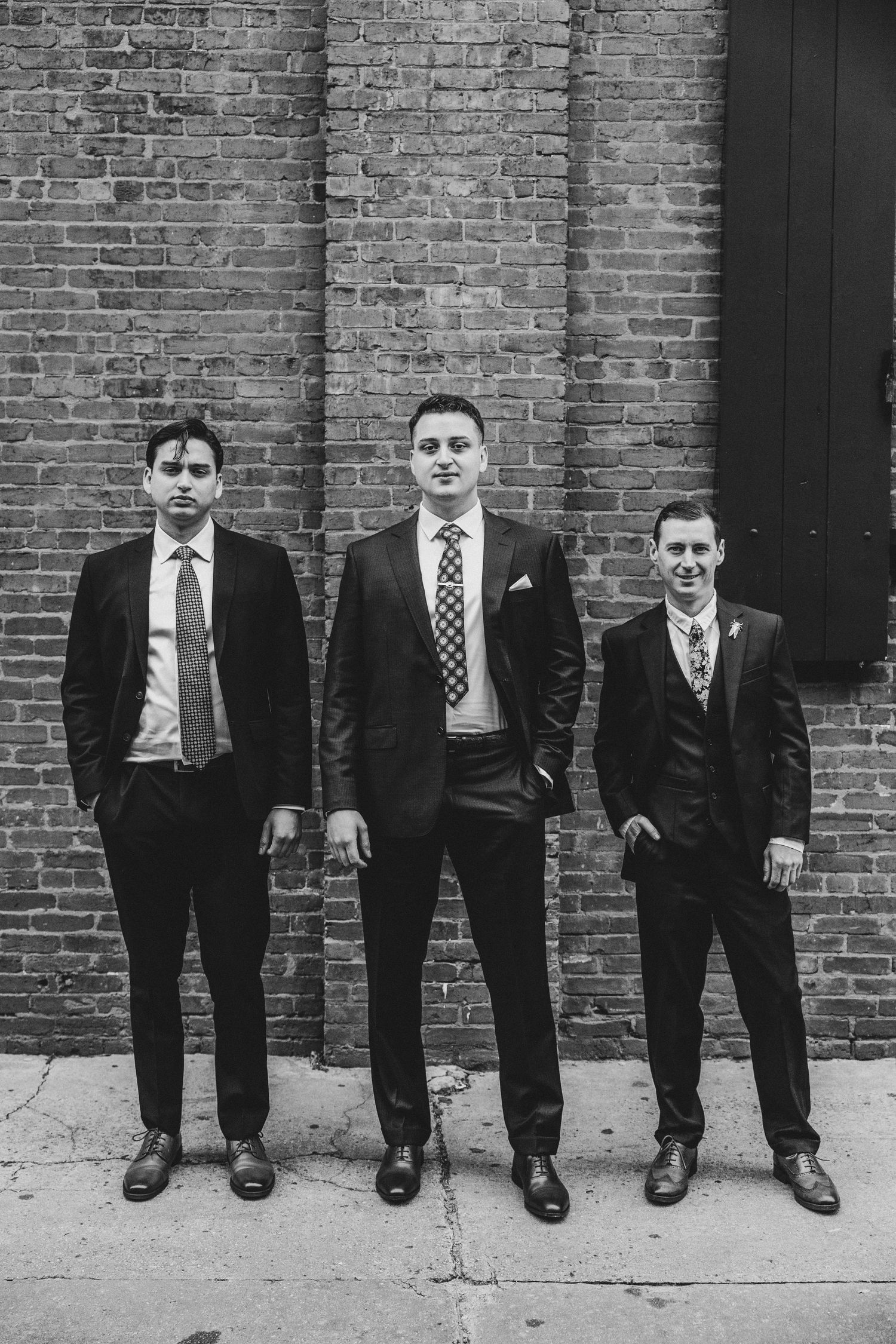 Three groomsmen in dark suits and ties pose confidently in front of a rustic brick wall in black and white.