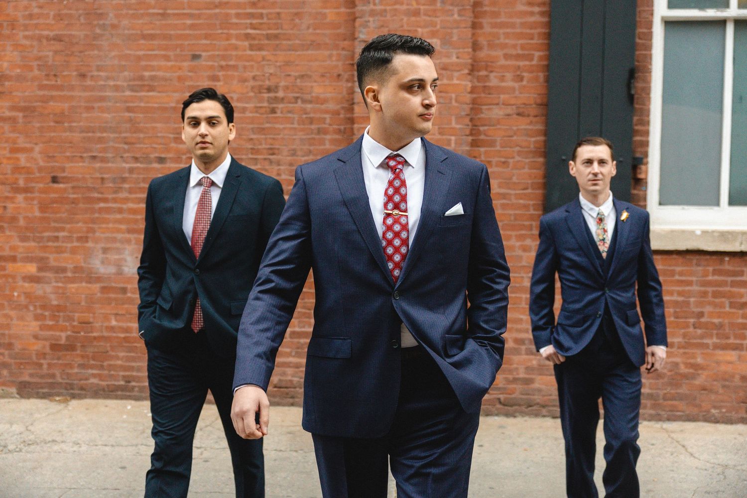 Three groomsmen in navy blue suits and red ties walking outdoors near a brick building.