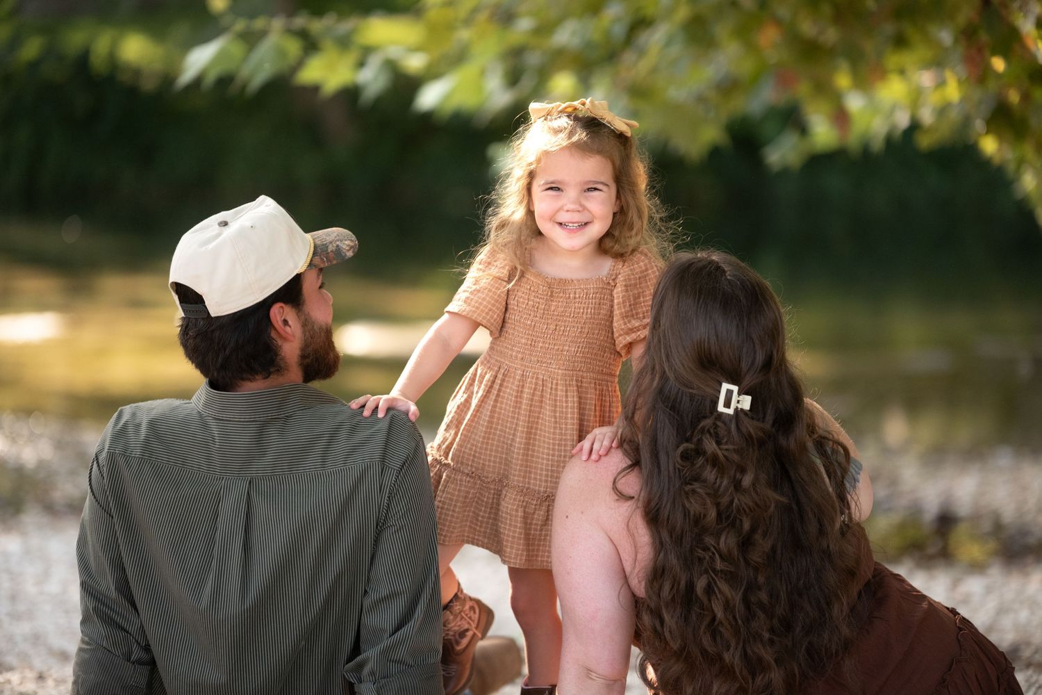 Parents with smiling toddler girl in tan dress outdoors near water with green trees in Salado, Texas