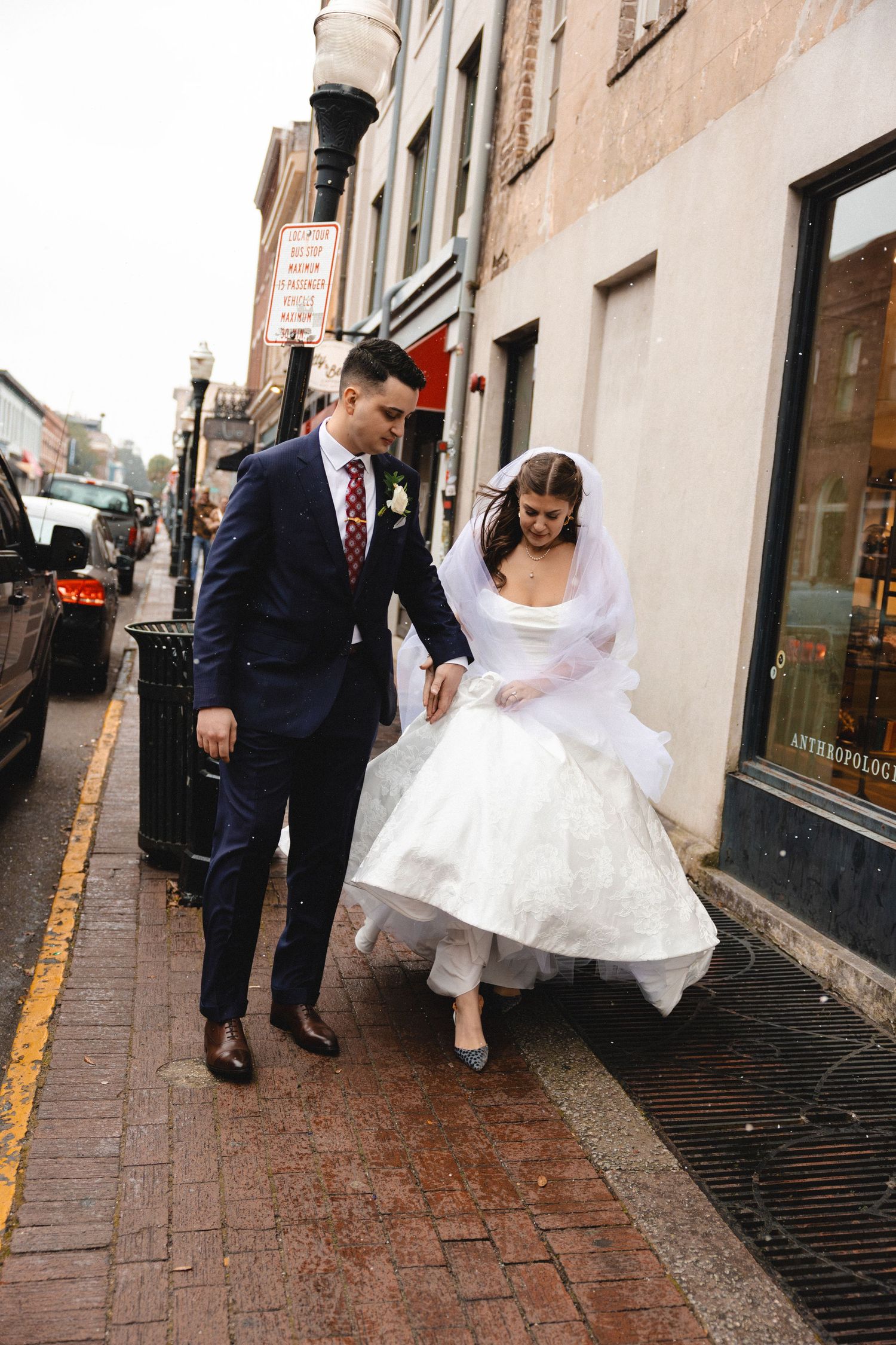 Bride in tea-length white dress and veil walks with groom in navy suit on a rainy city sidewalk.
