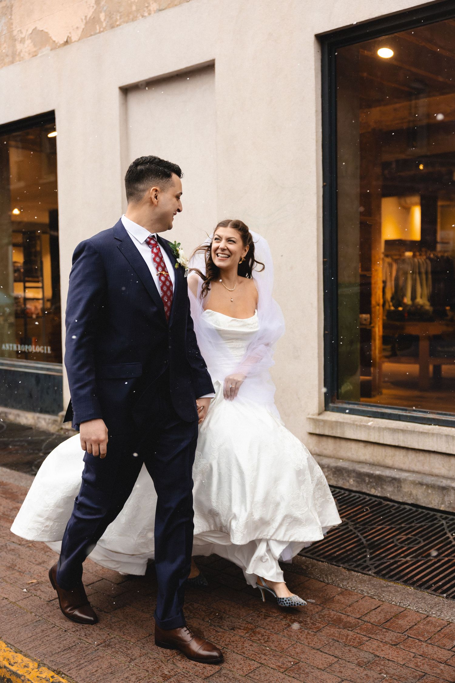 Bride and groom walking and laughing together on a city sidewalk, bride in white gown with veil, groom in navy suit.
