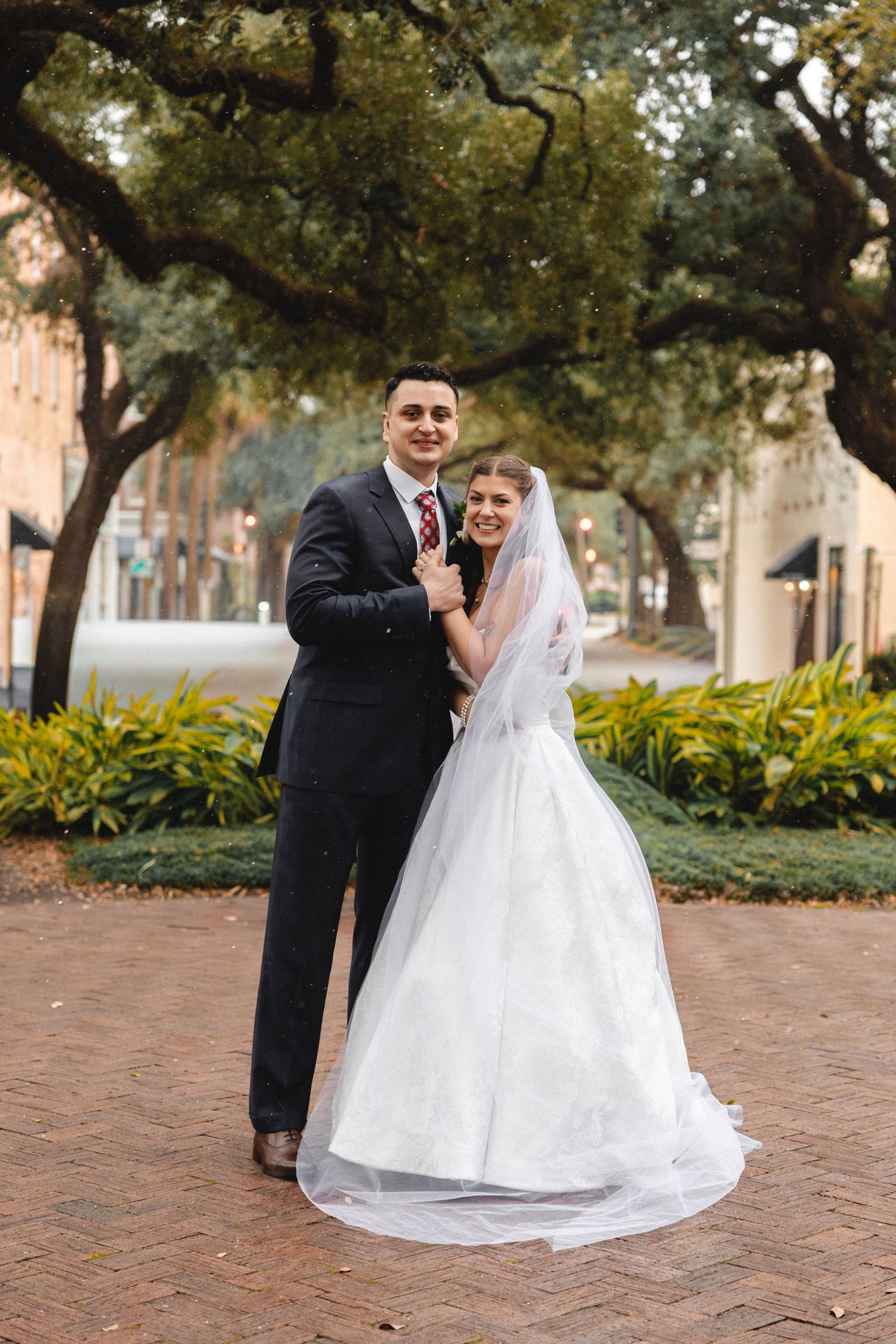 Bride and groom posing outdoors on brick path under oak trees, bride in white ballgown with veil, groom in dark suit.