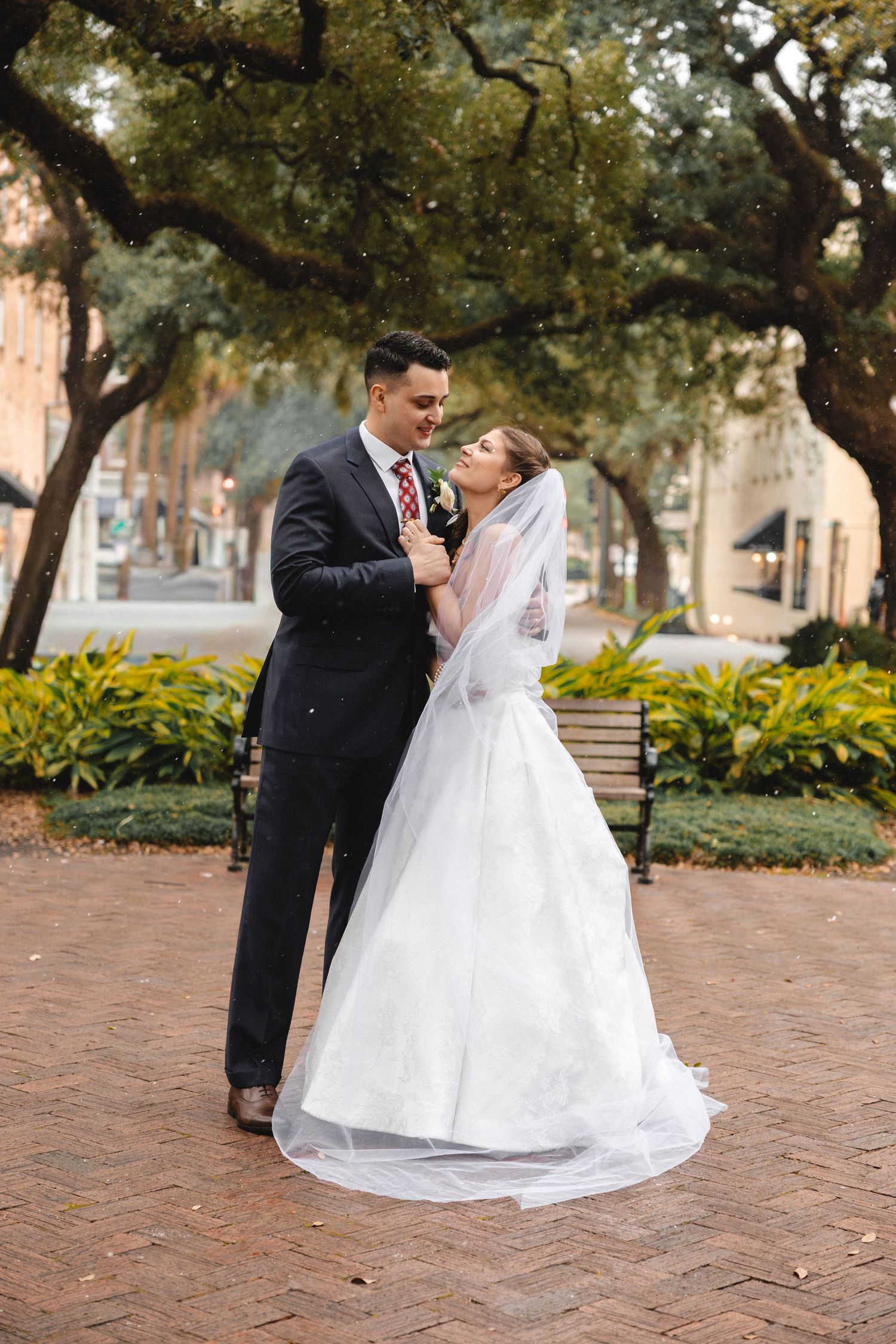 Bride and groom share a romantic embrace on a brick plaza surrounded by lush trees.