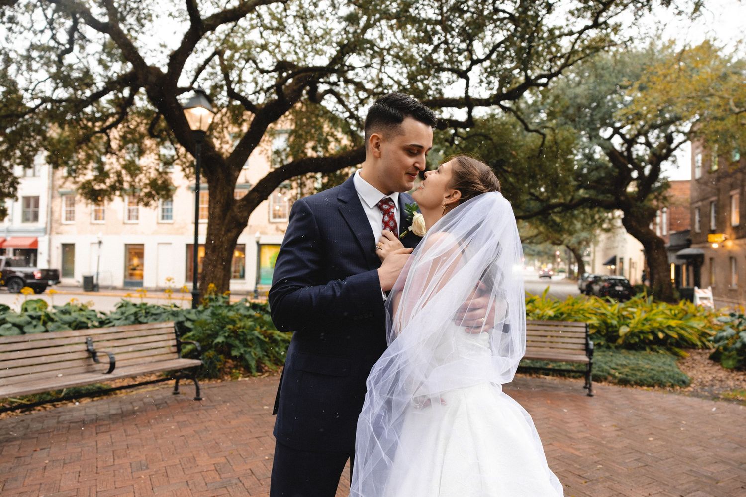 A bride and groom share a romantic moment in a scenic outdoor plaza, surrounded by lush trees and park benches.