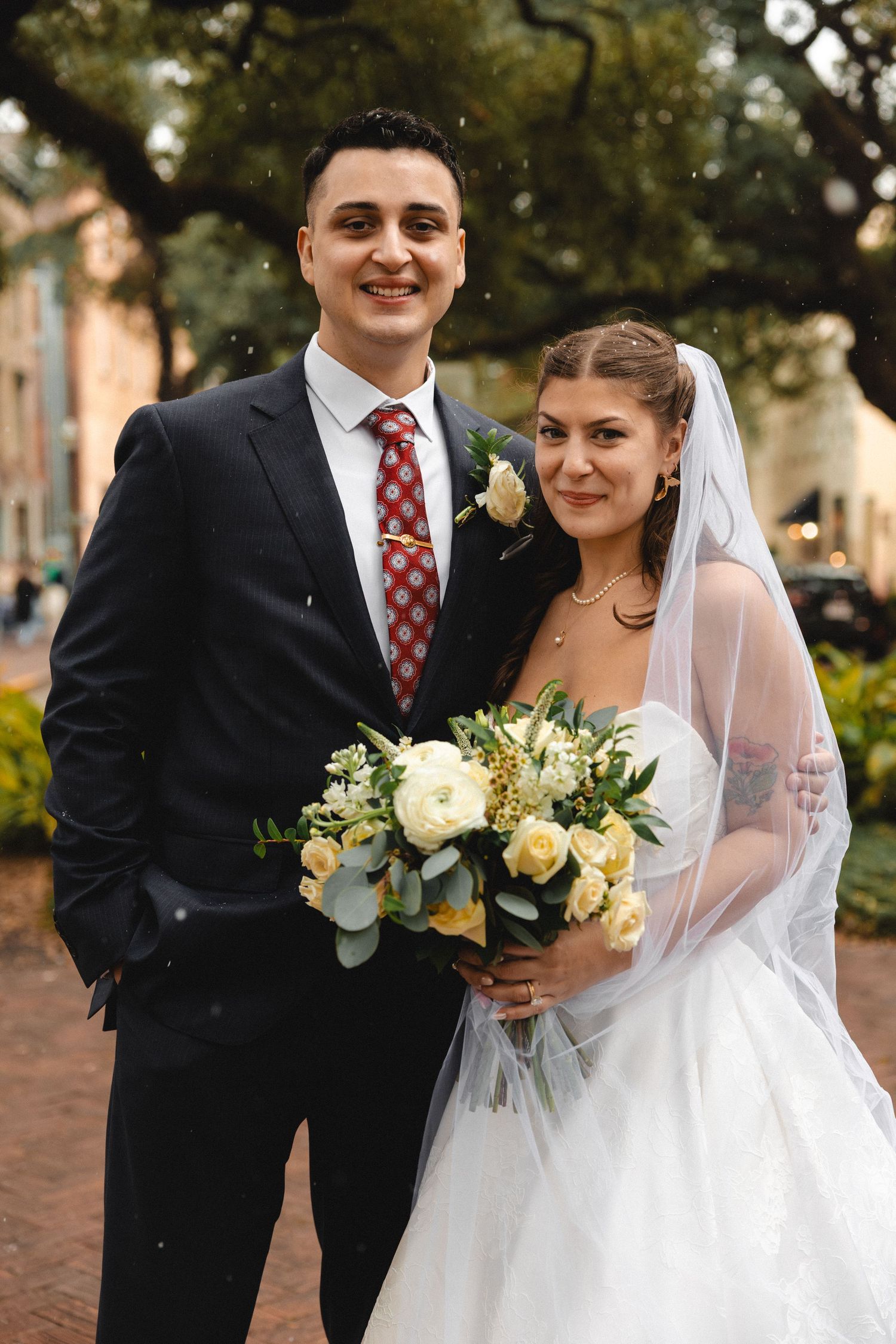 Bride and groom posing outdoors on their wedding day, she holds a white and yellow bouquet while wearing a veil.