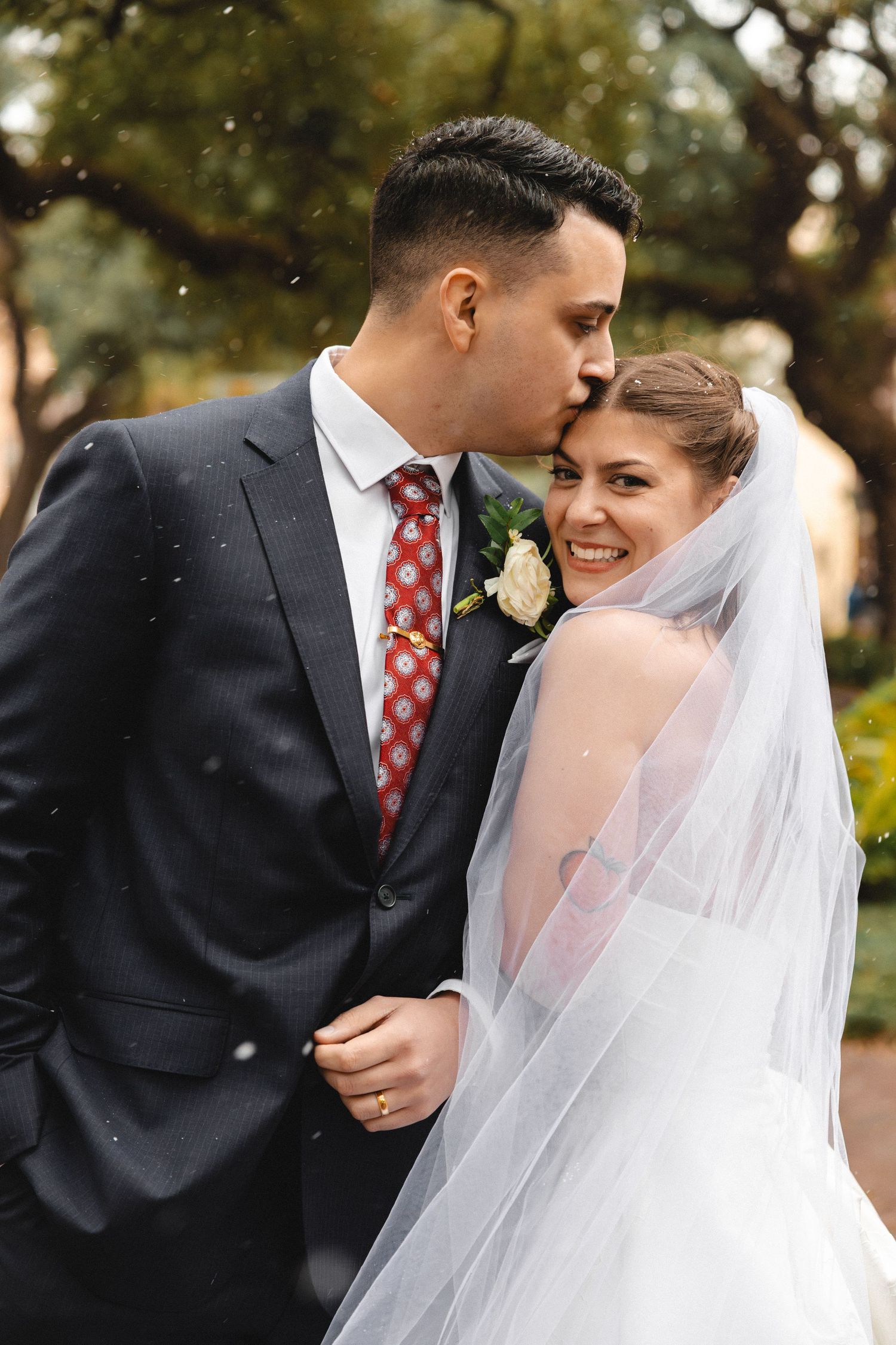Groom in navy suit kisses bride on forehead as she smiles, wearing white wedding gown and veil outdoors.