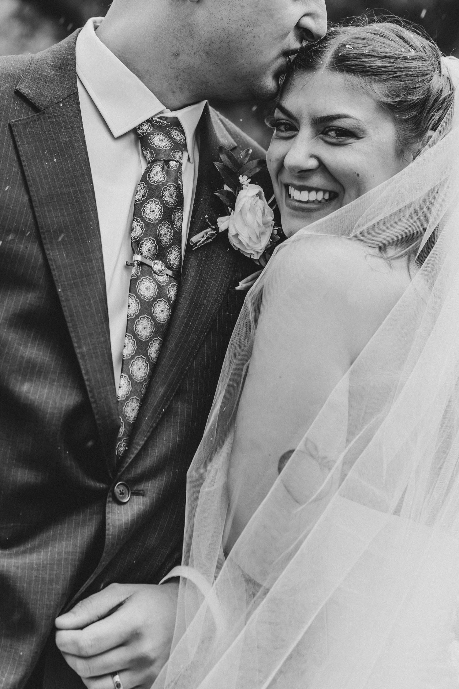 Black and white photo of a groom kissing bride on forehead as she smiles joyfully, veil draped over shoulder.