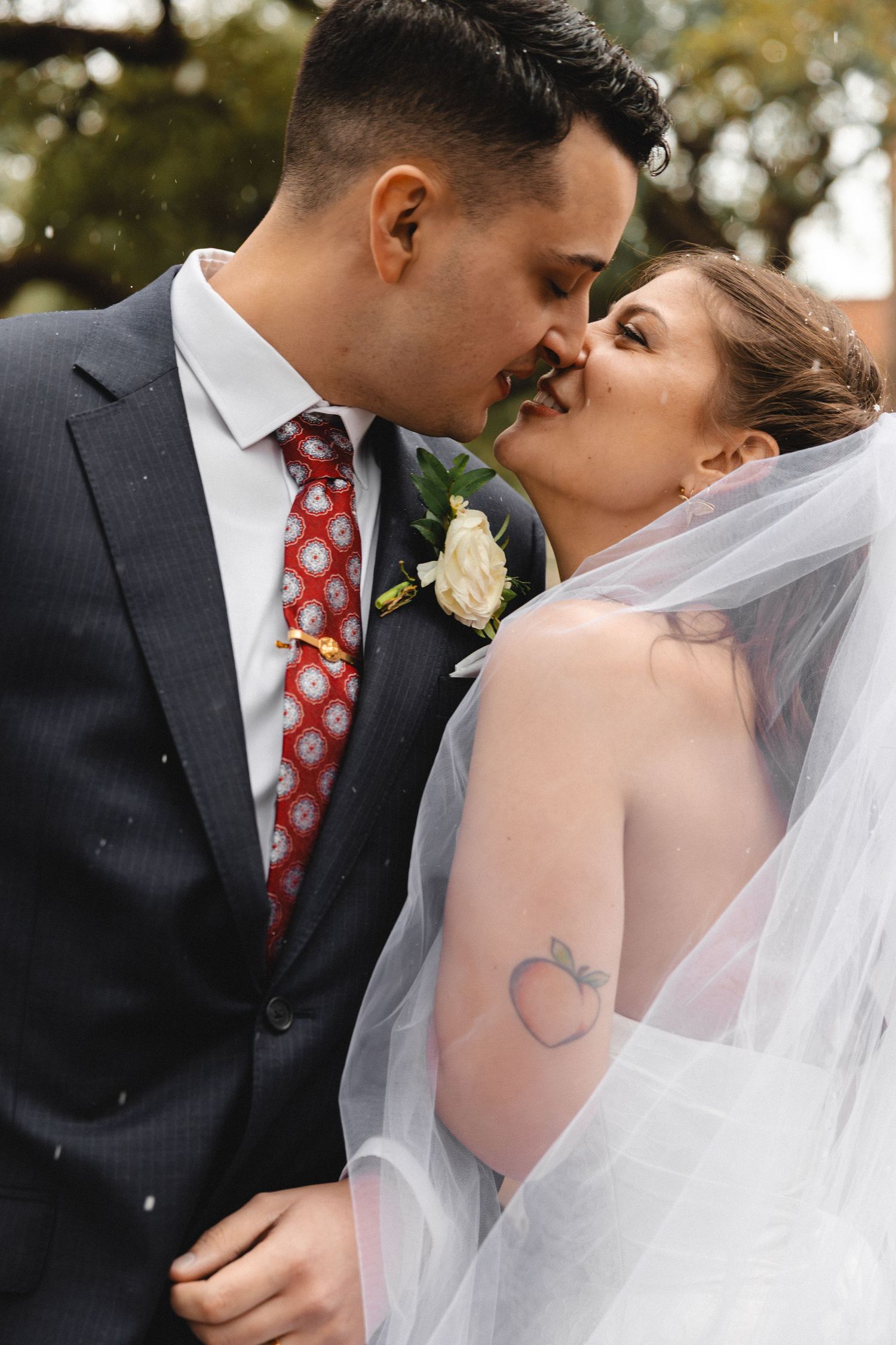 A bride and groom share a romantic kiss, with the bride wearing a veil and backless gown with a peach tattoo.