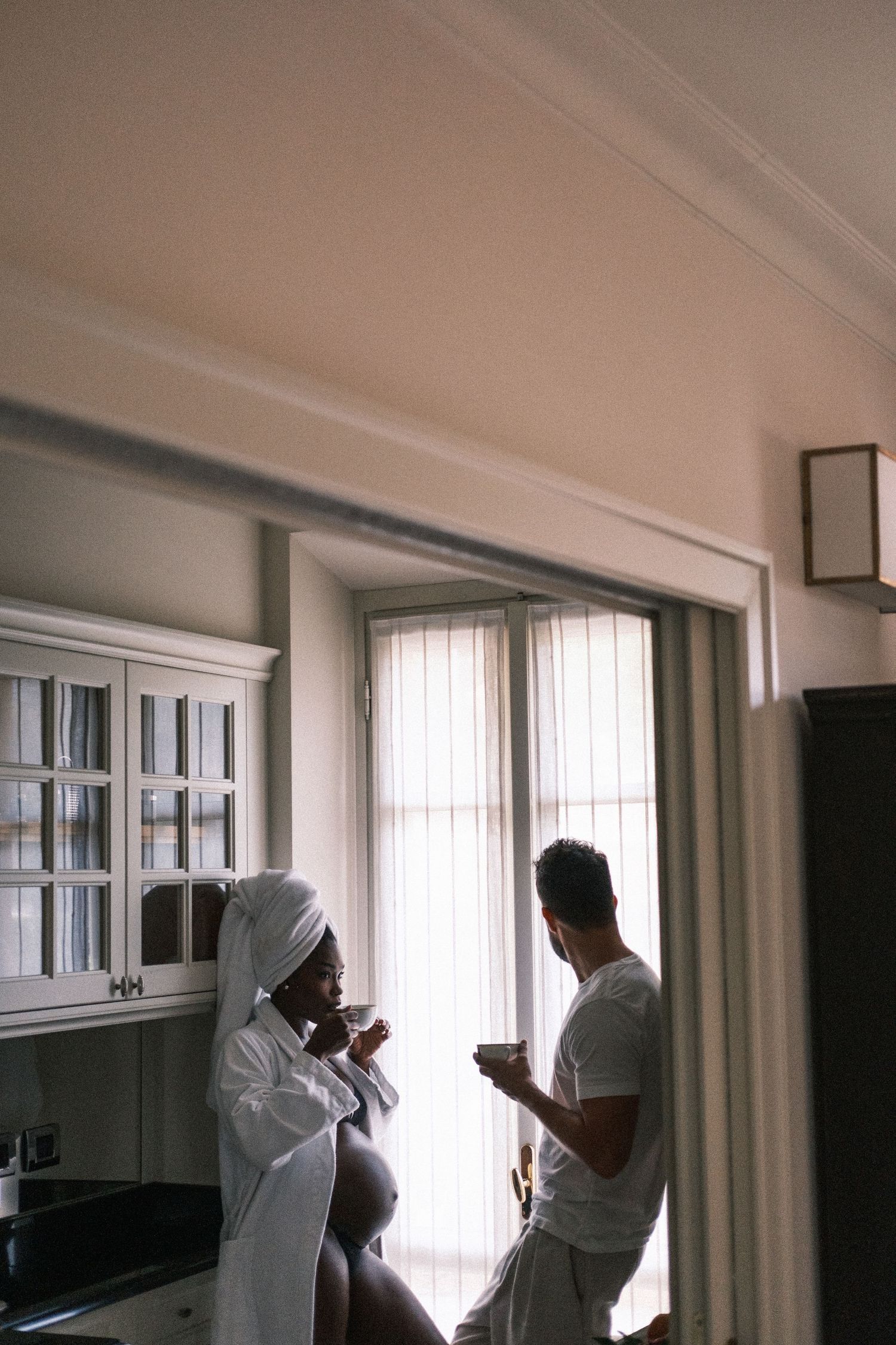 Couple in robes enjoying morning coffee in a bright kitchen, woman with towel on head, man by window.