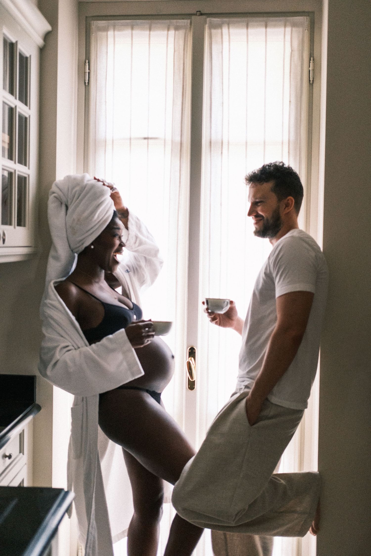 Pregnant woman in robe and towel turban shares morning coffee with smiling man by sunlit window.