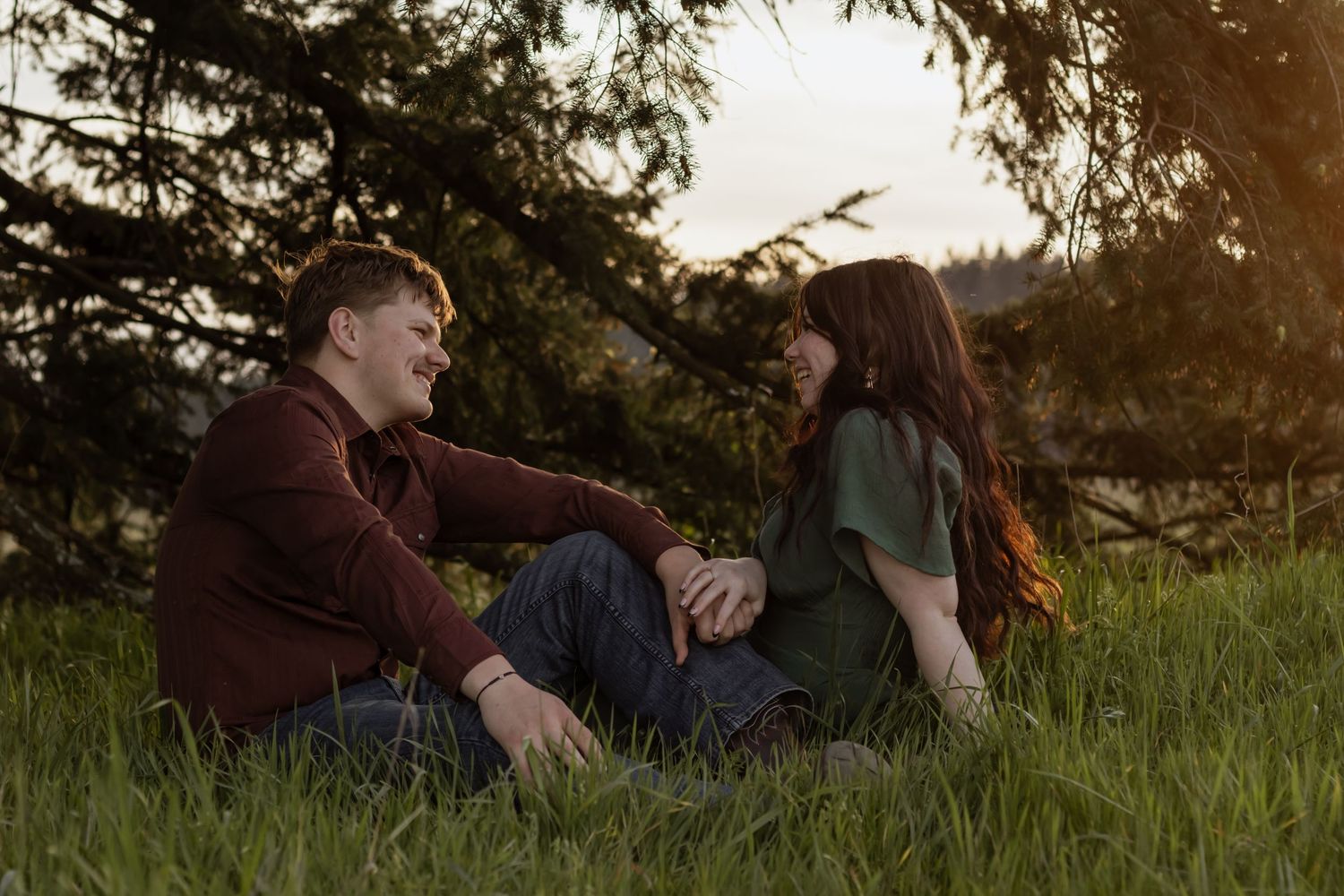 Couple sitting together on green grass under pine trees during golden hour sunset light.