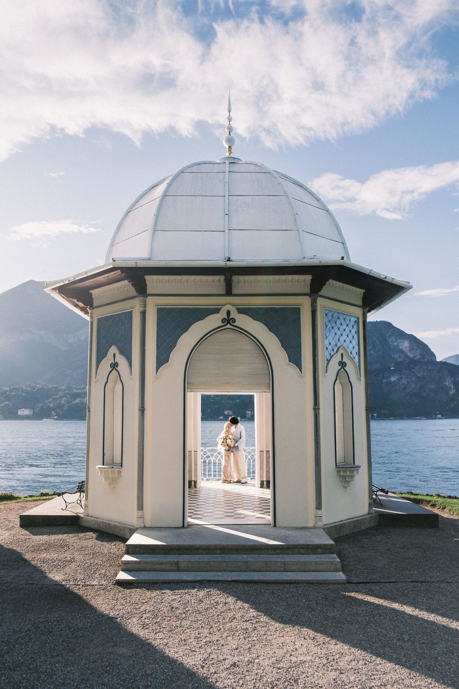 Elegant domed pavilion with arched doorways overlooking Lake Como, Italy, with mountains and blue sky backdrop.