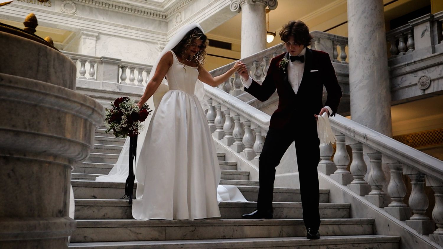 Bride and groom descend grand marble staircase in elegant wedding attire, bride holding bouquet in classic venue.