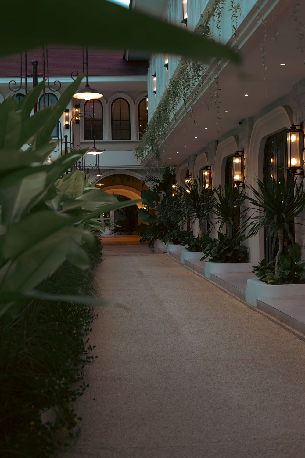 Elegant indoor corridor with lush tropical plants, arched doorways, and warm pendant lighting in a luxury hotel.