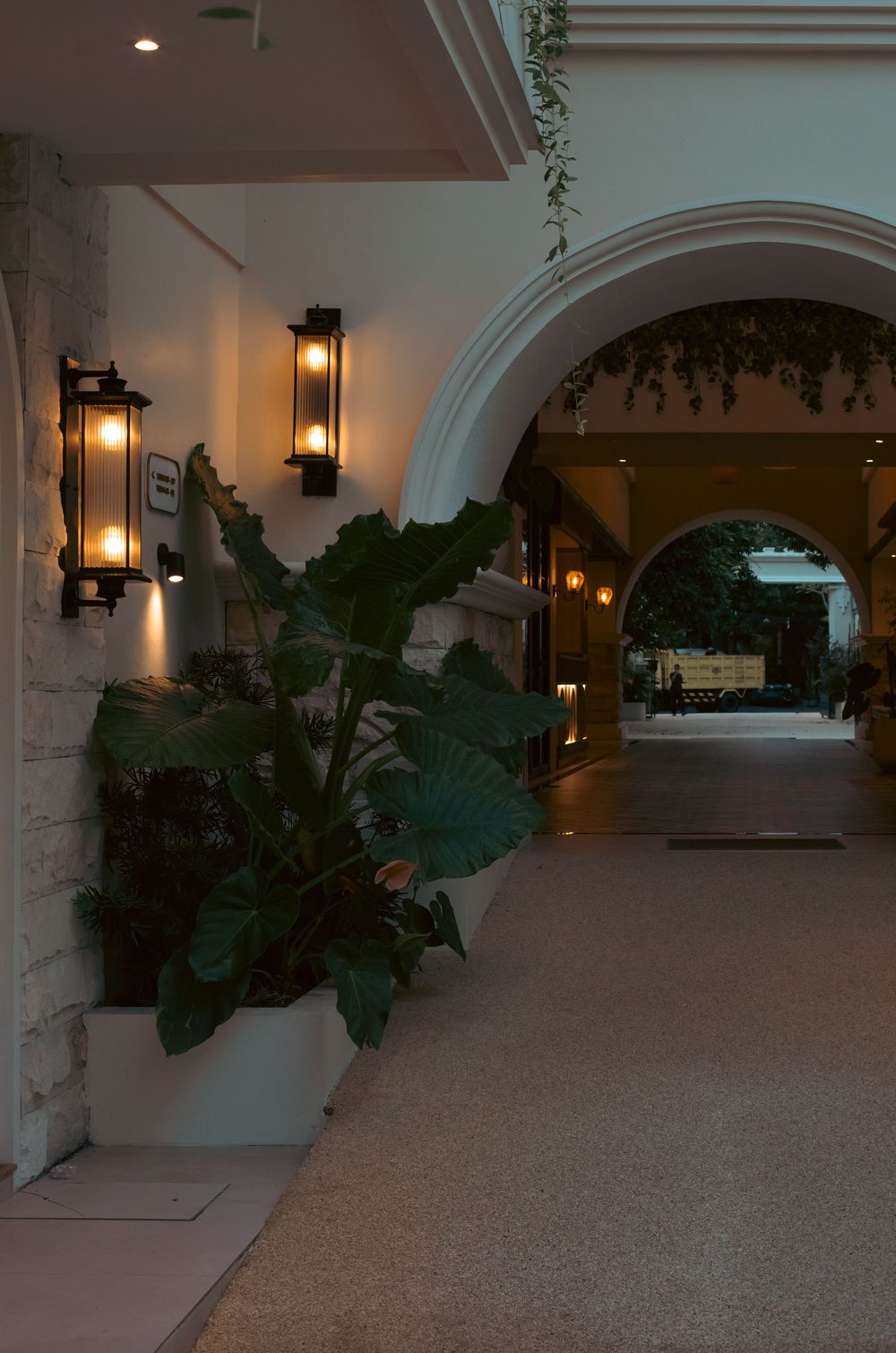 Elegant hotel corridor with arched doorways, wall lanterns, and tropical potted plants in warm evening lighting.