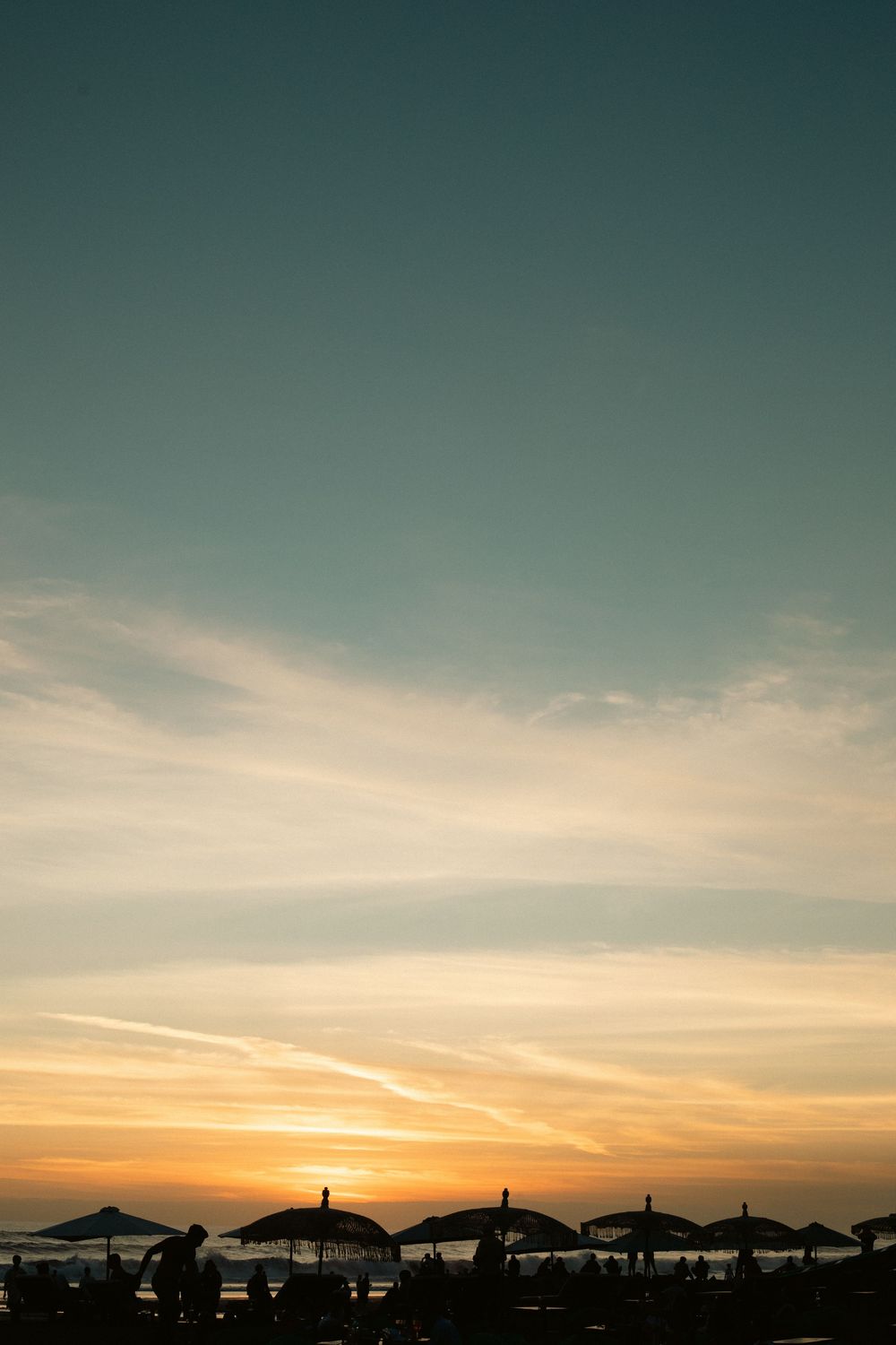Silhouettes of beach umbrellas against a vibrant sunset sky with golden orange hues over the ocean horizon.