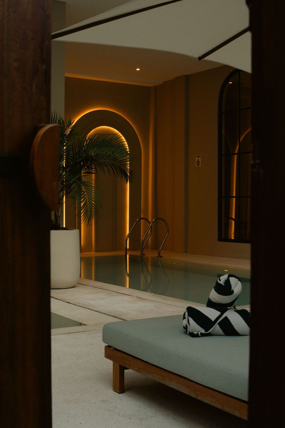 Elegant indoor pool with arched walls, warm amber lighting, tropical plant, and a daybed with striped cushions.