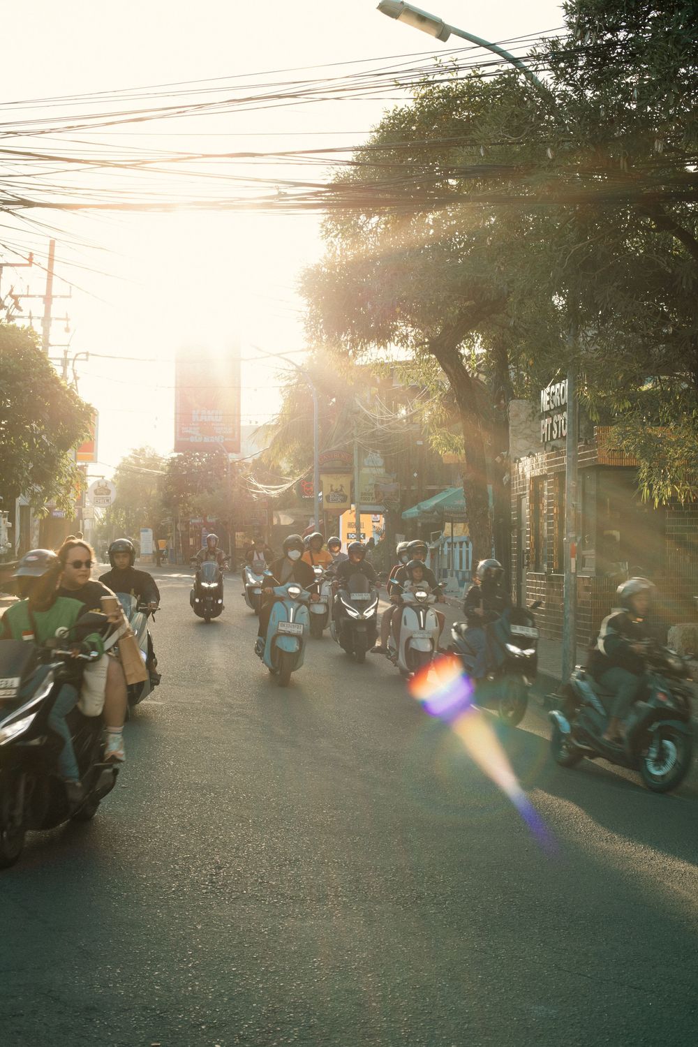 Motorcyclists riding through a busy Southeast Asian street at golden hour with sun flare and overhead wires.
