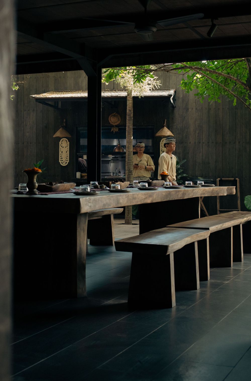 Two chefs prepare food at a rustic wooden outdoor kitchen with bench seating under a dark pavilion.