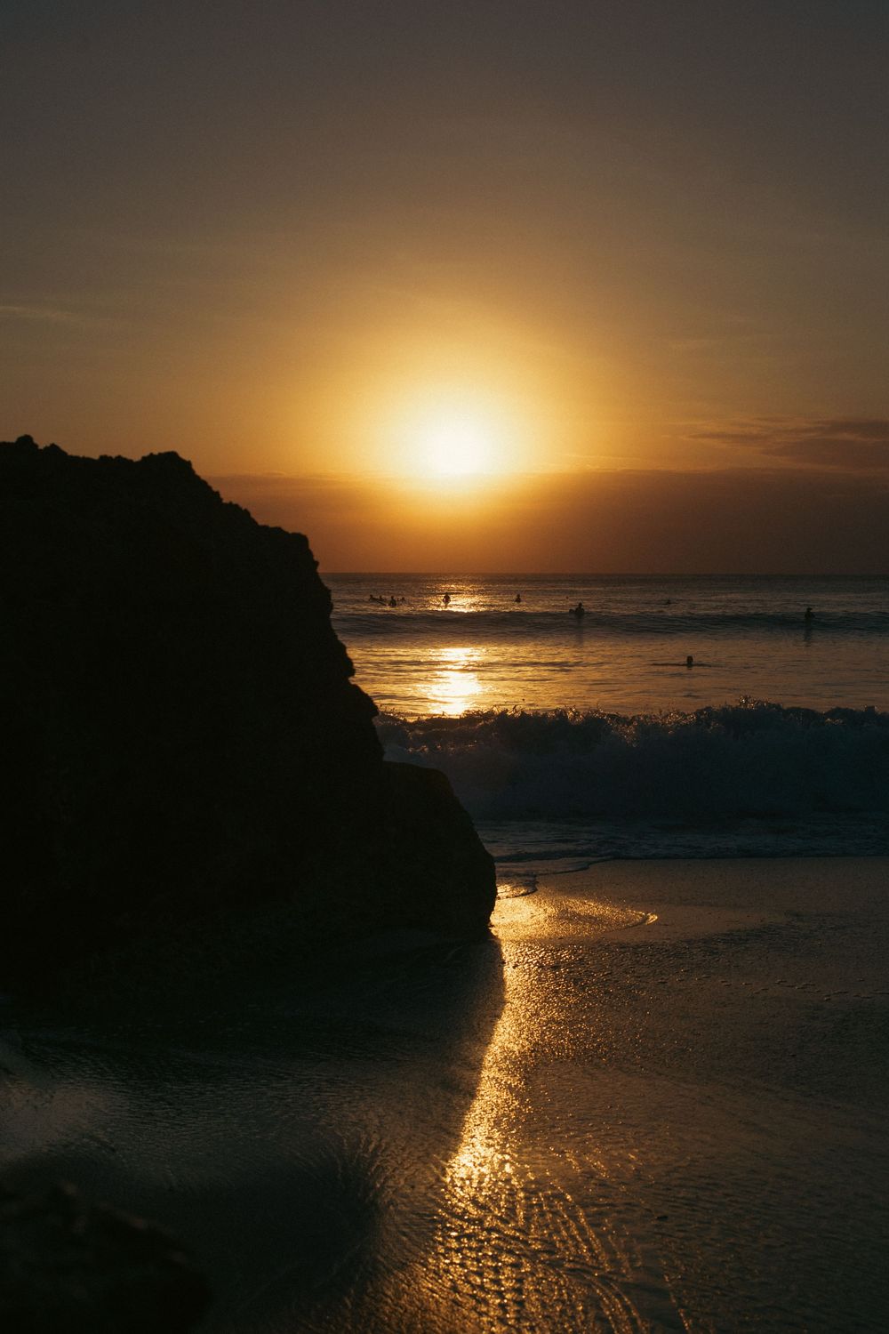 Golden sunset over ocean waves with silhouetted rock formation and surfers in the water at a scenic beach.