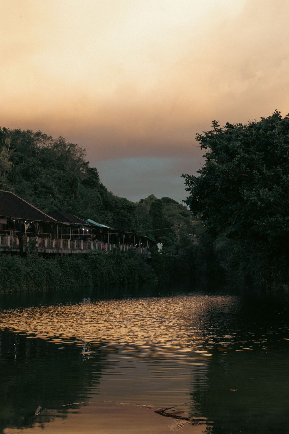 Golden sunset reflects on a calm river canal surrounded by lush green trees and a rustic wooden building at dusk.