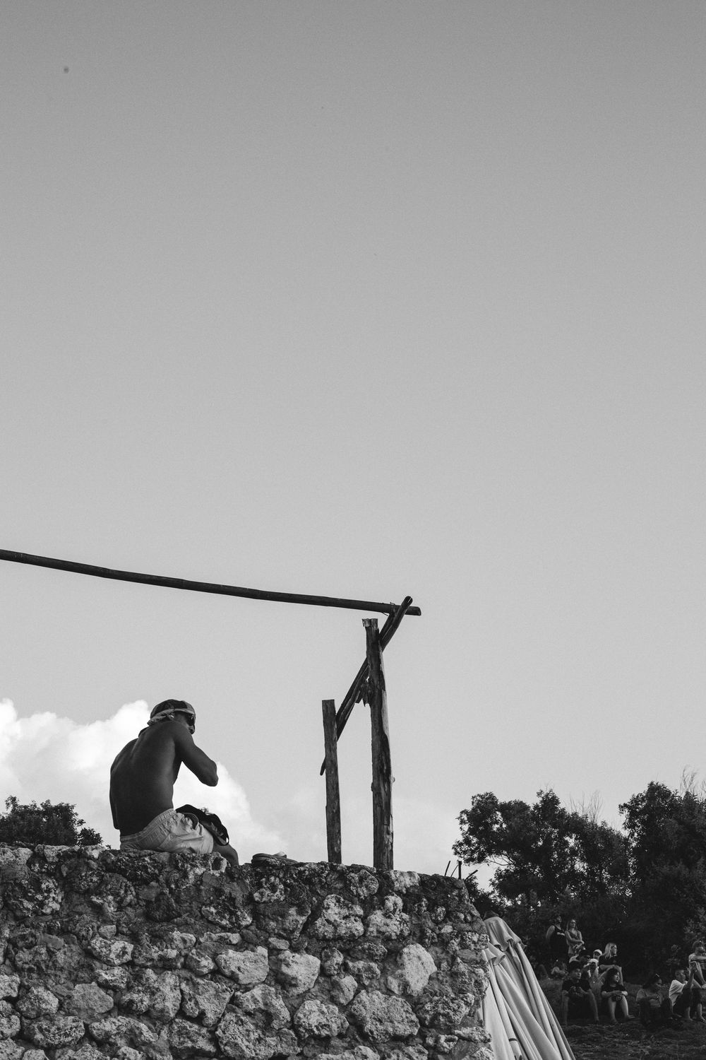 Black and white photo of a person sitting on a stone wall beside a wooden post with rope against a cloudy sky.