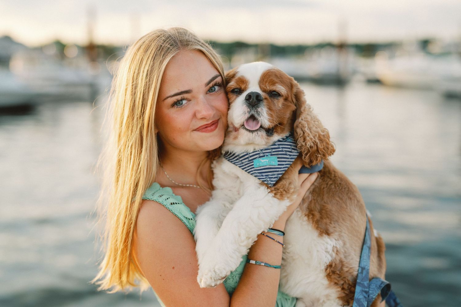 Young blonde woman smiling and holding a Cavalier King Charles Spaniel dog near a marina at sunset.