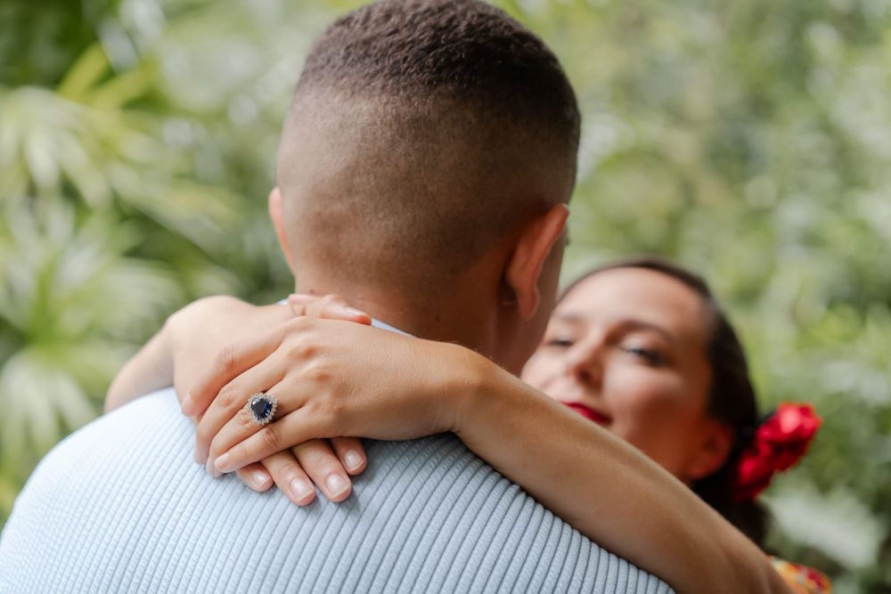 Woman with a red flower in her hair hugs a man, her hand showing a sapphire engagement ring, against lush green tropical foliage.