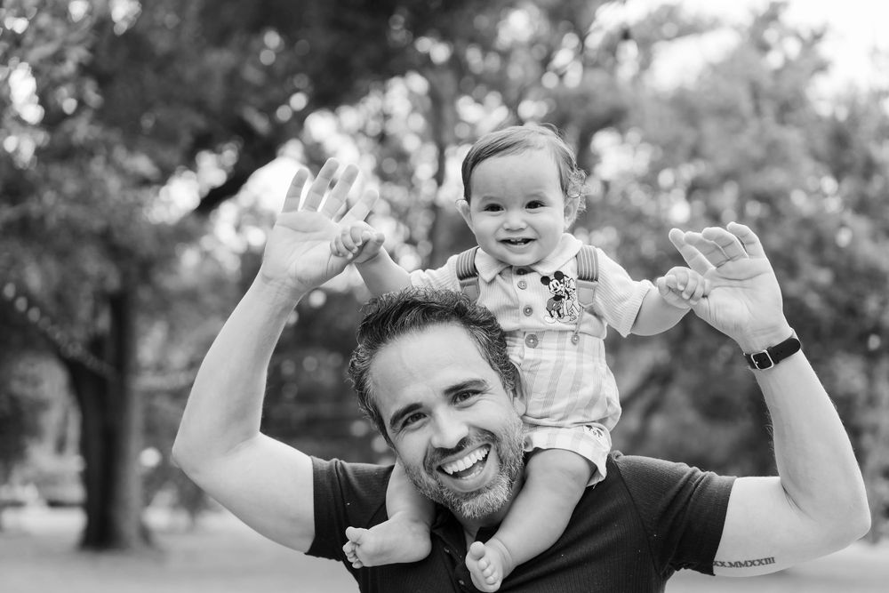 Smiling bearded father holds his laughing baby on his shoulders as both raise their hands in a joyful black and white outdoor portrait.