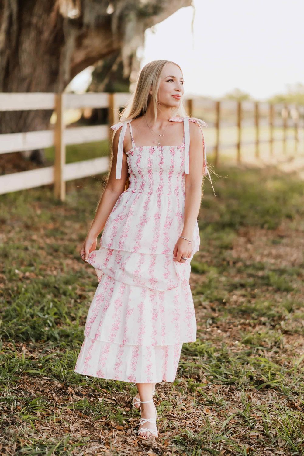 Woman in a pink floral tiered smocked midi dress with tie shoulder straps and white sandals walks along a wooden fence in a grassy field.
