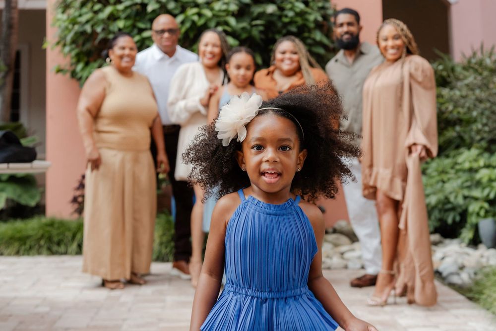 Smiling young girl in a blue dress with a flower headband stands outdoors while her happy multigenerational family poses behind her.
