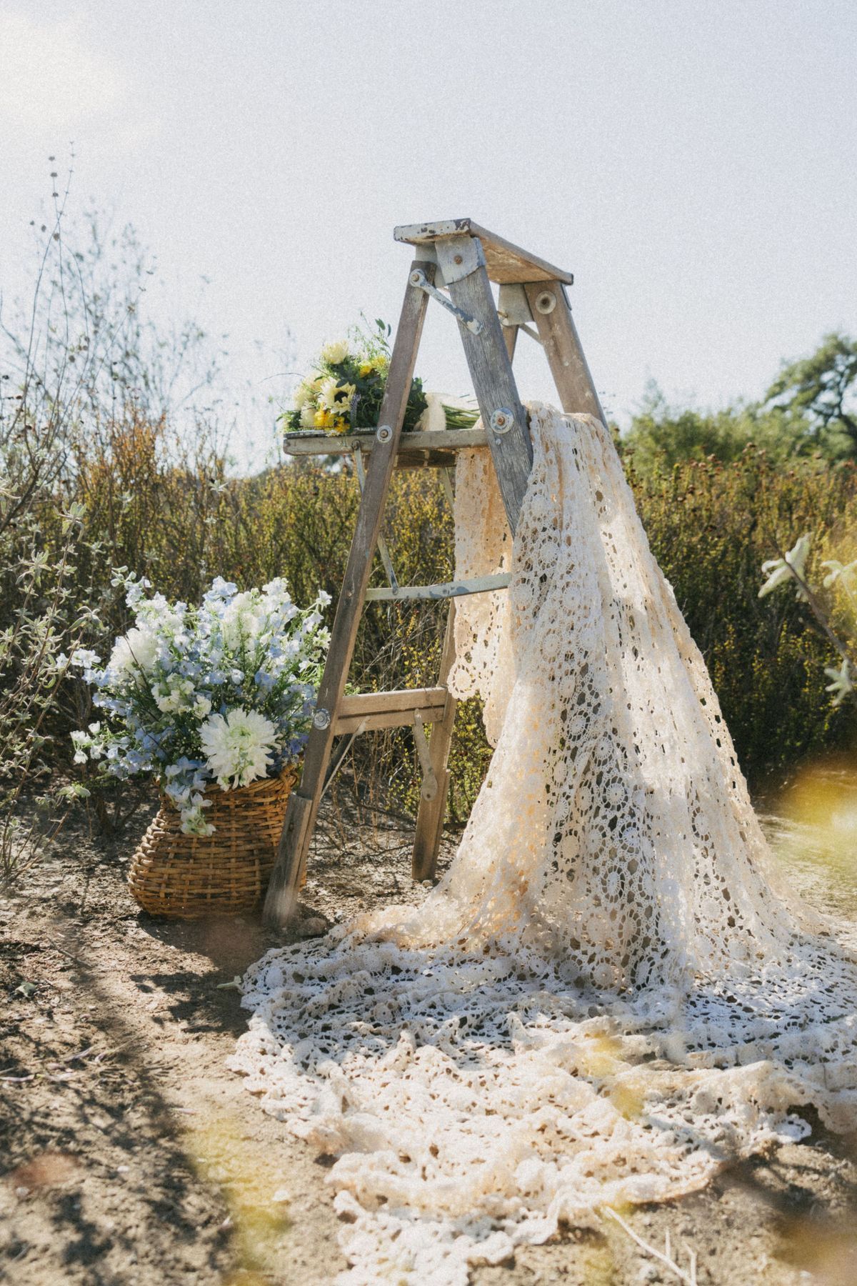 Rustic wooden ladder with lace fabric and floral basket arrangement in a natural outdoor setting.