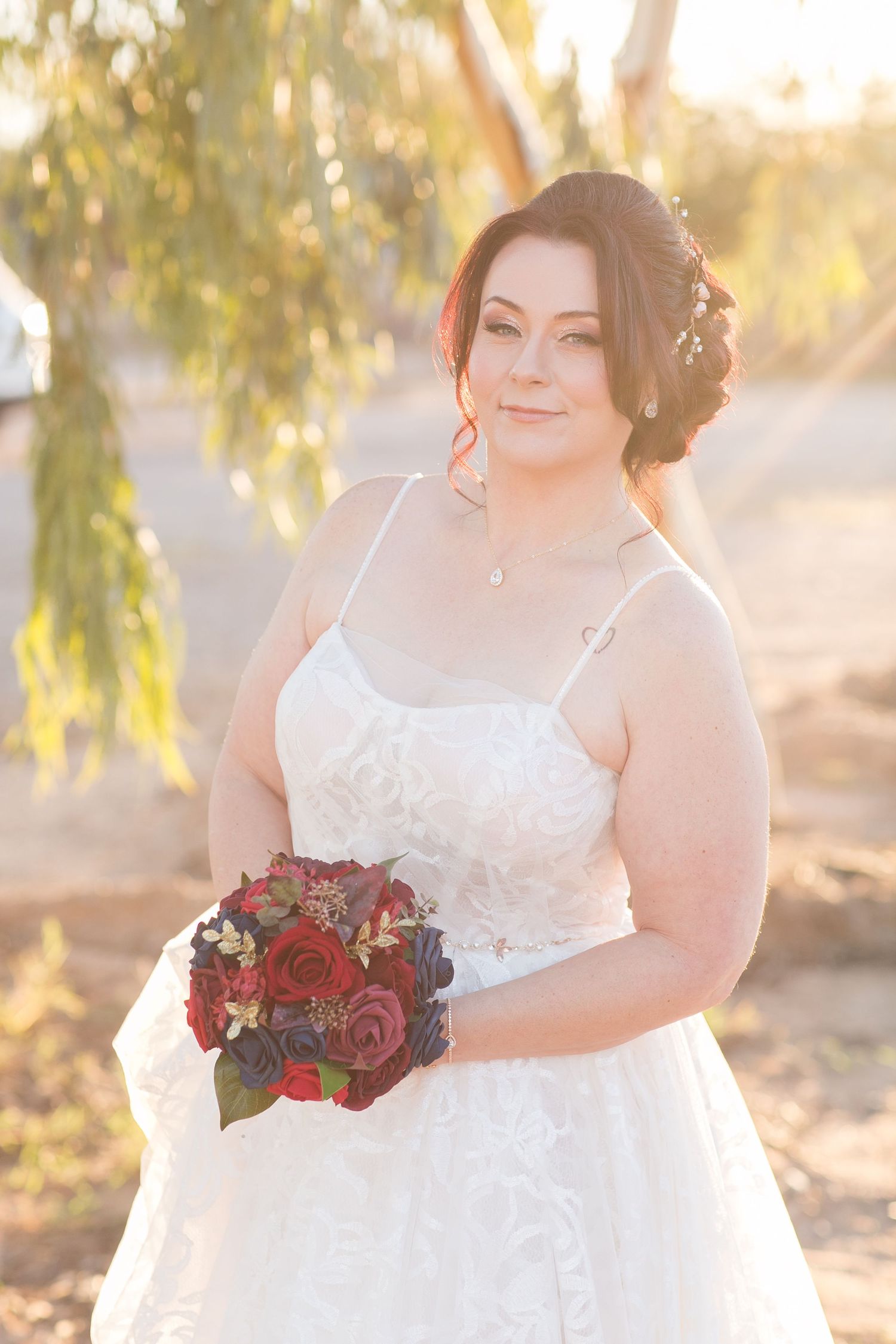 Bride in white lace gown holding dark red rose bouquet outdoors in golden hour light.