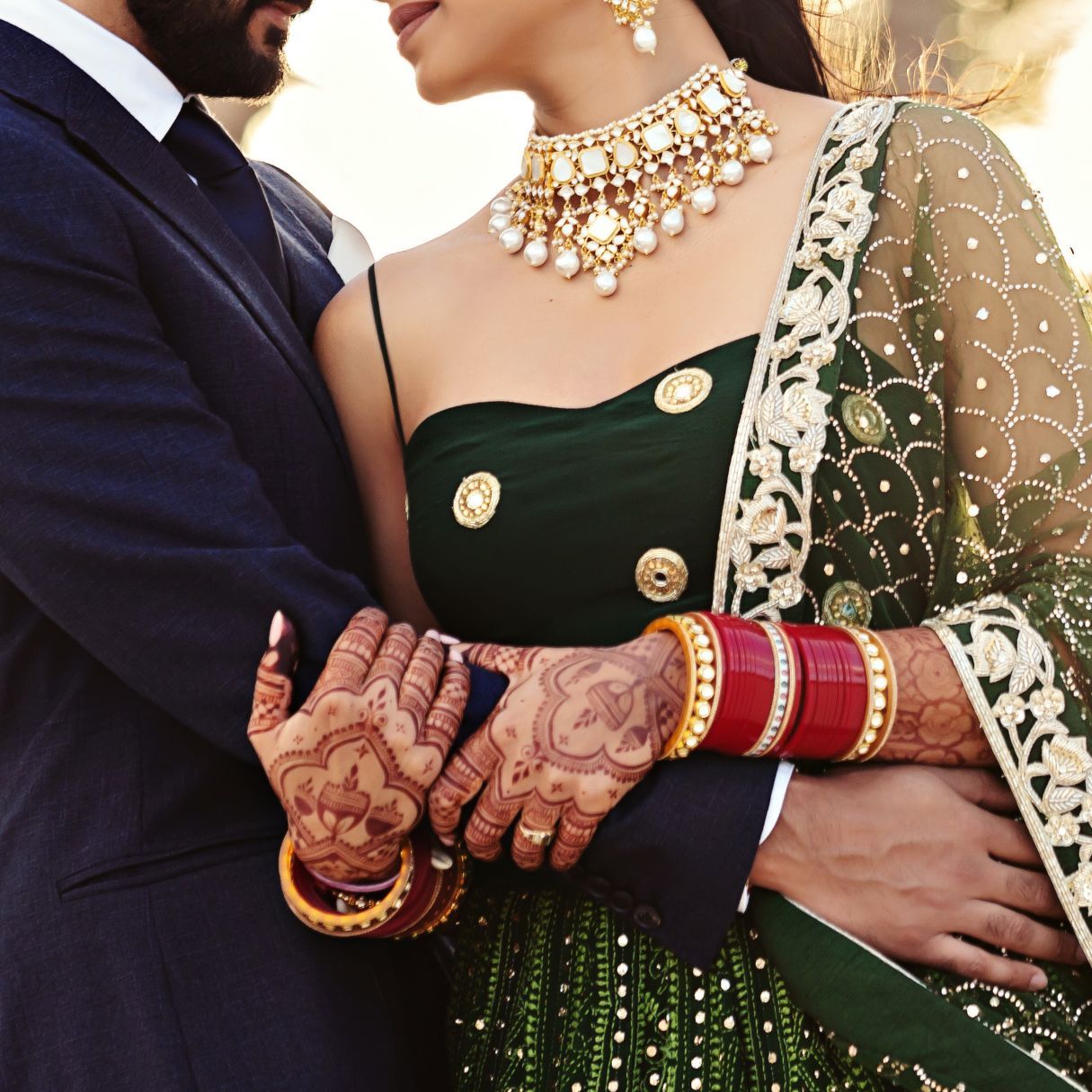 Indian bride and groom in traditional attire, bride wearing green saree with gold jewelry and mehndi, holding hands tenderly.