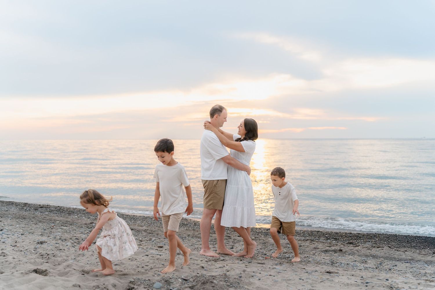 Family of five enjoying a sunset beach family photo session, parents embracing while children play at Lake Erie, in Mentor, Ohio.