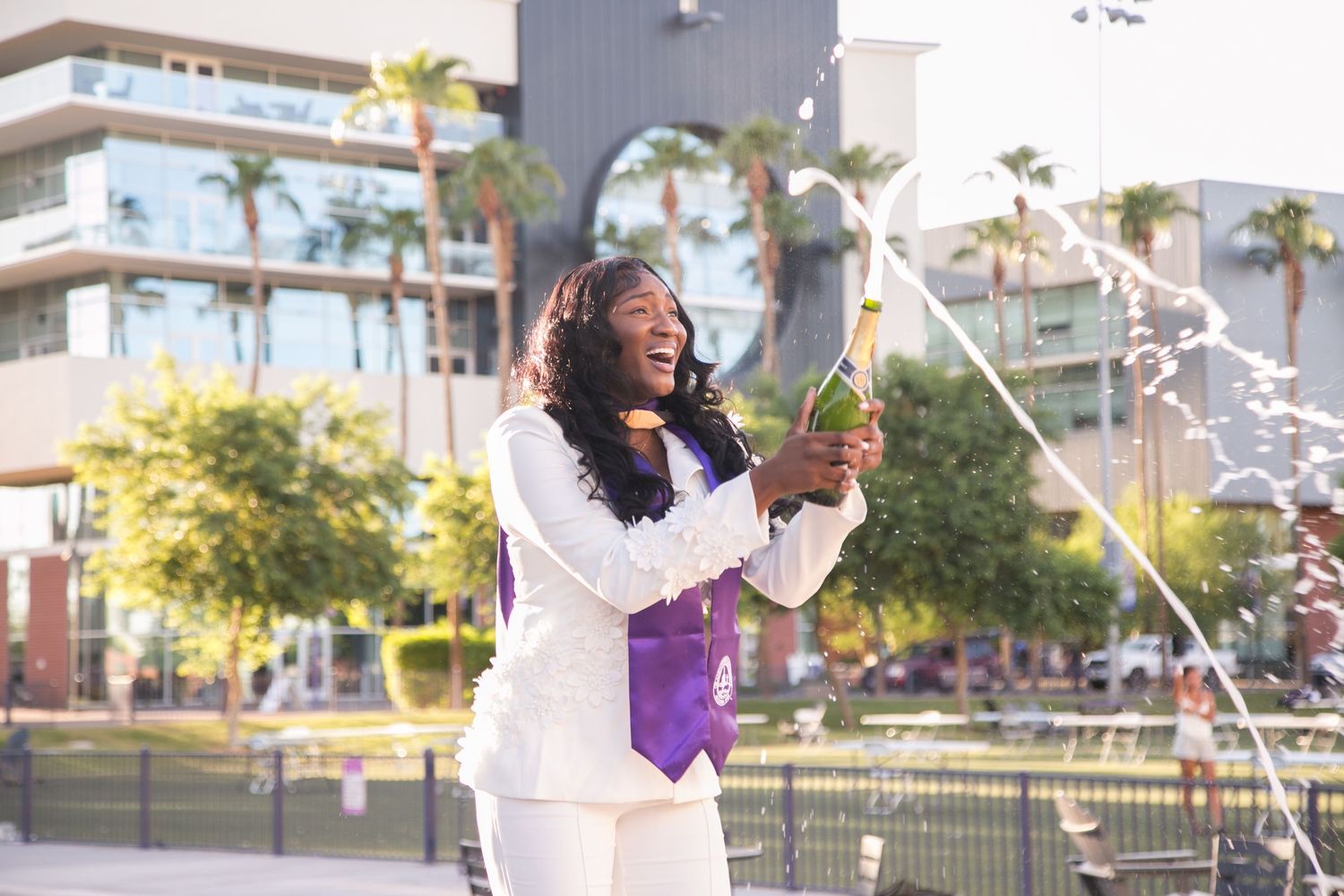 Graduate in white suit pops champagne bottle outdoors, celebrating academic achievement with purple sash.